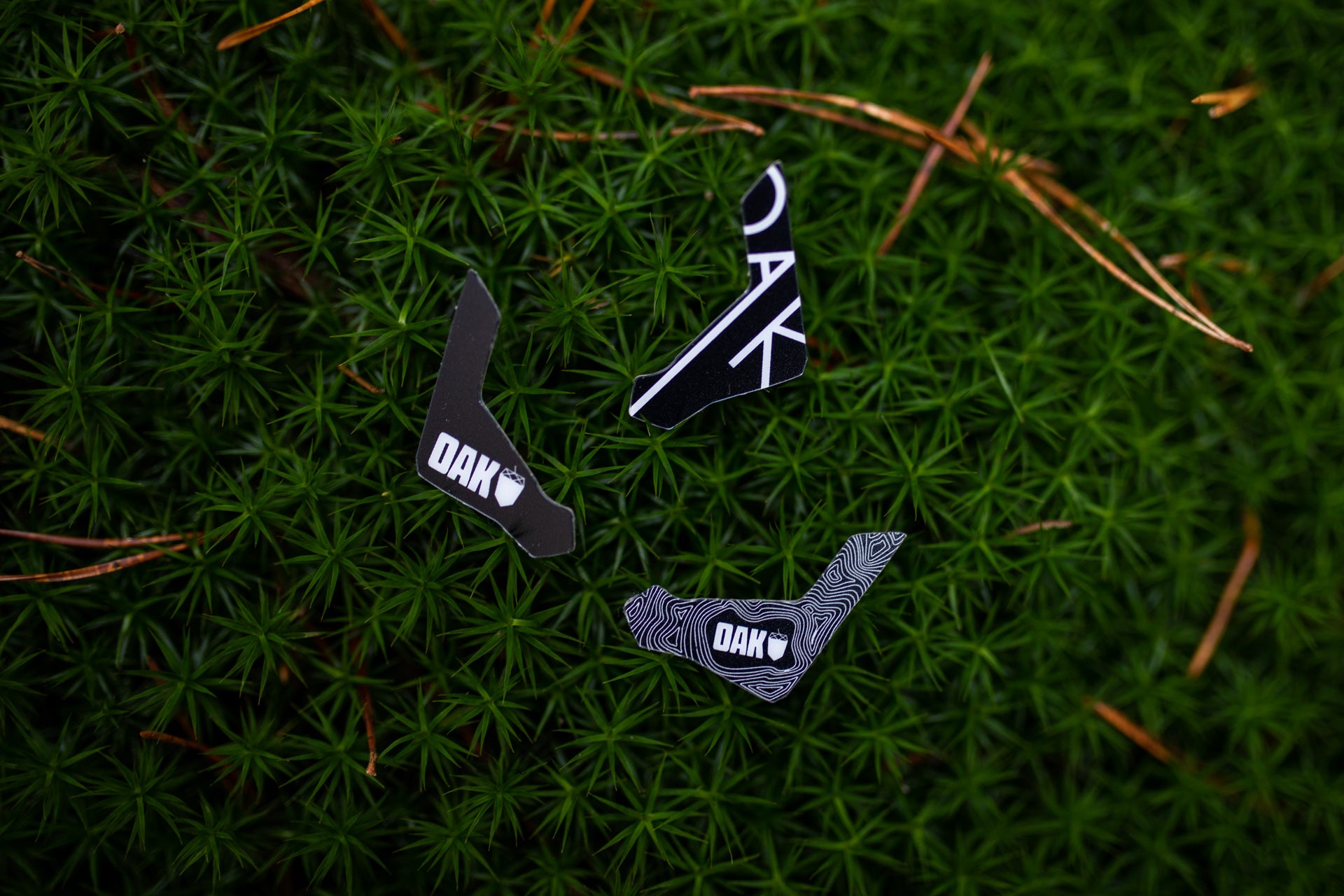 Three black OAK decals with white lettering on green moss and pine needles, showcasing outdoor durability.