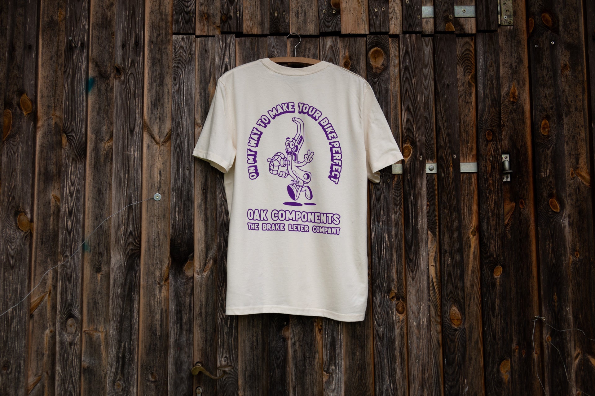 Cream T-shirt with purple cartoon brake lever and OAK Components slogan on wooden wall.