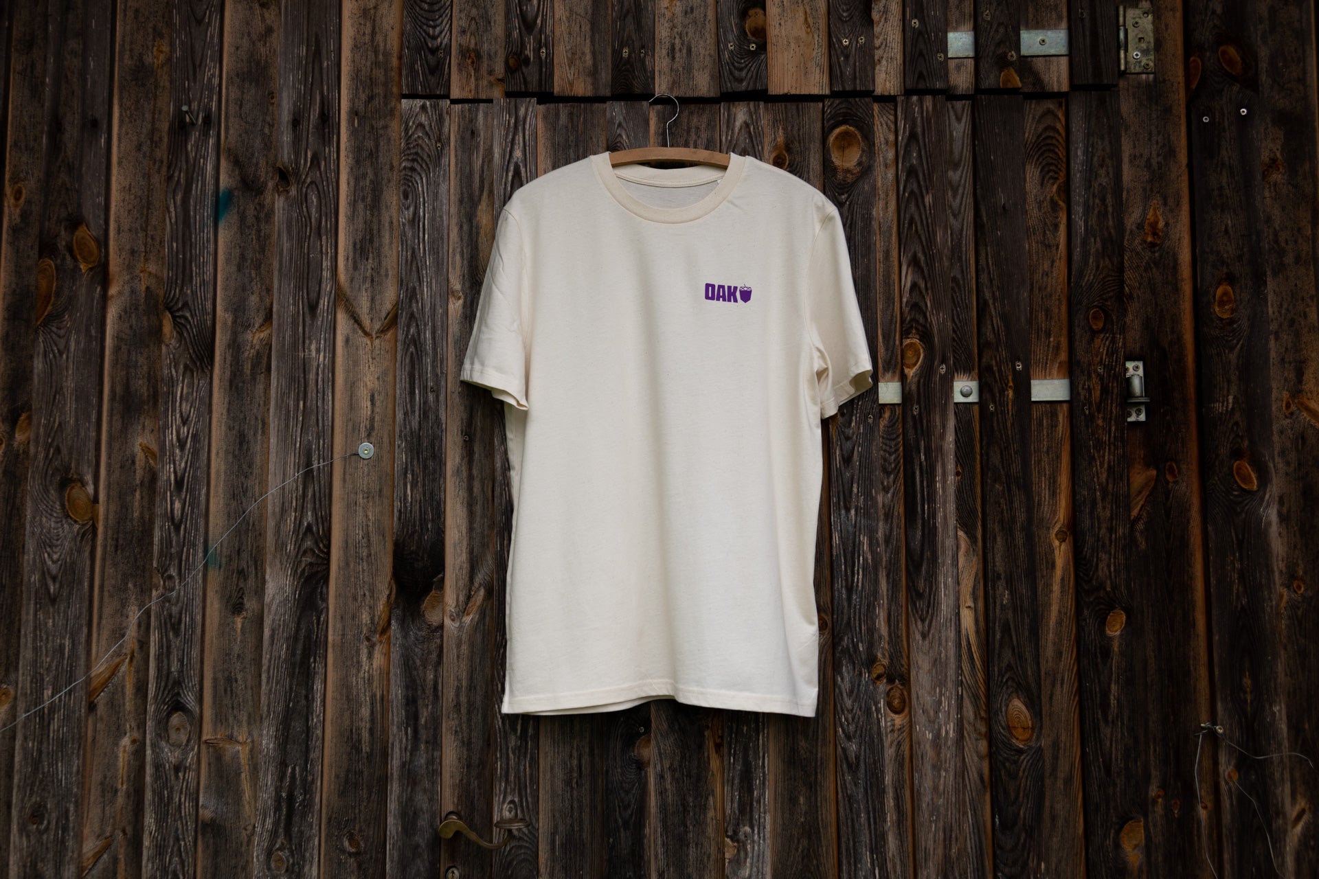 Off-white t-shirt with purple DAK logo and acorn symbol on left chest, on wooden hanger against dark wood wall
