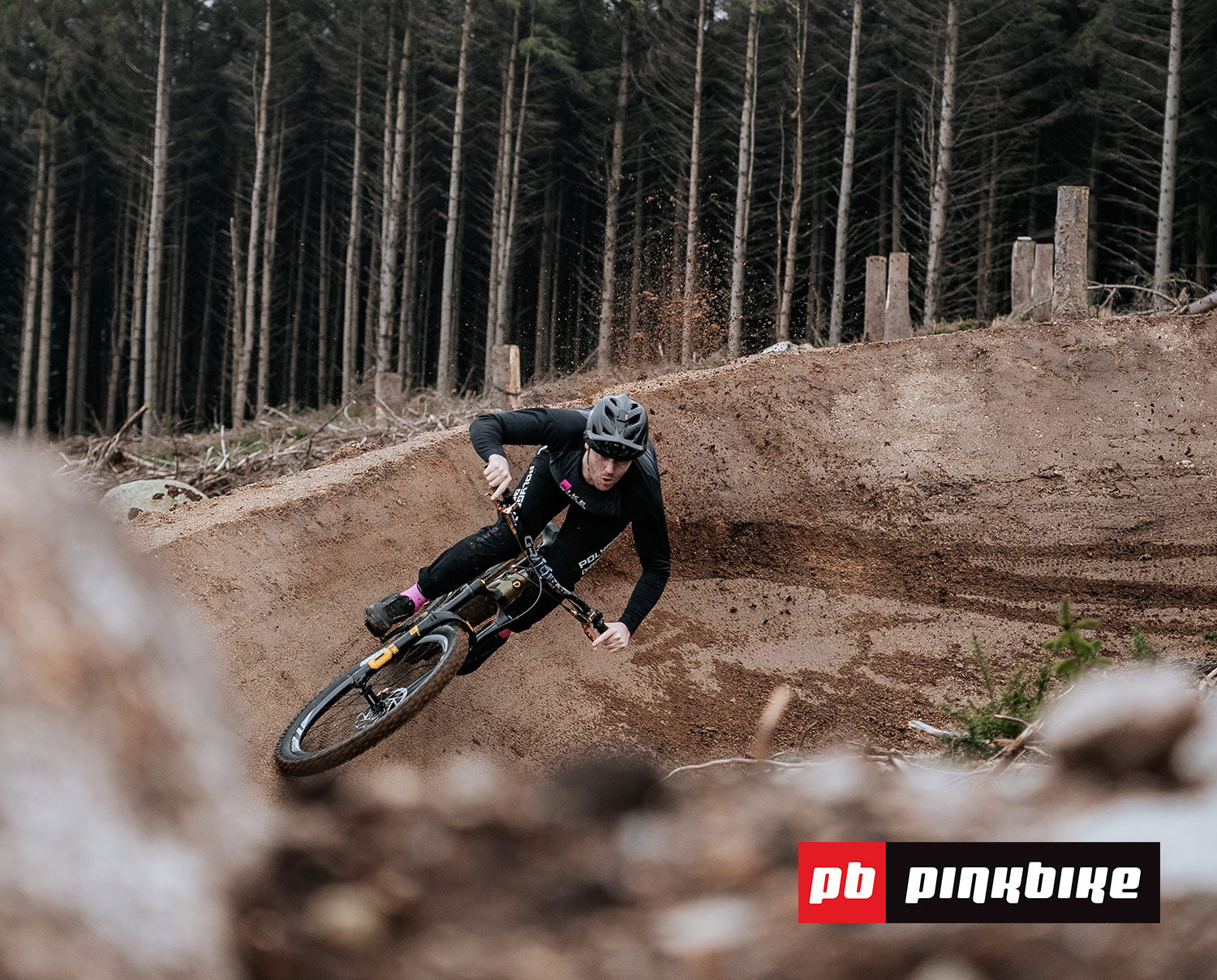 Cyclist in black and pink gear leaning into a dirt berm on a forest trail with leafless trees