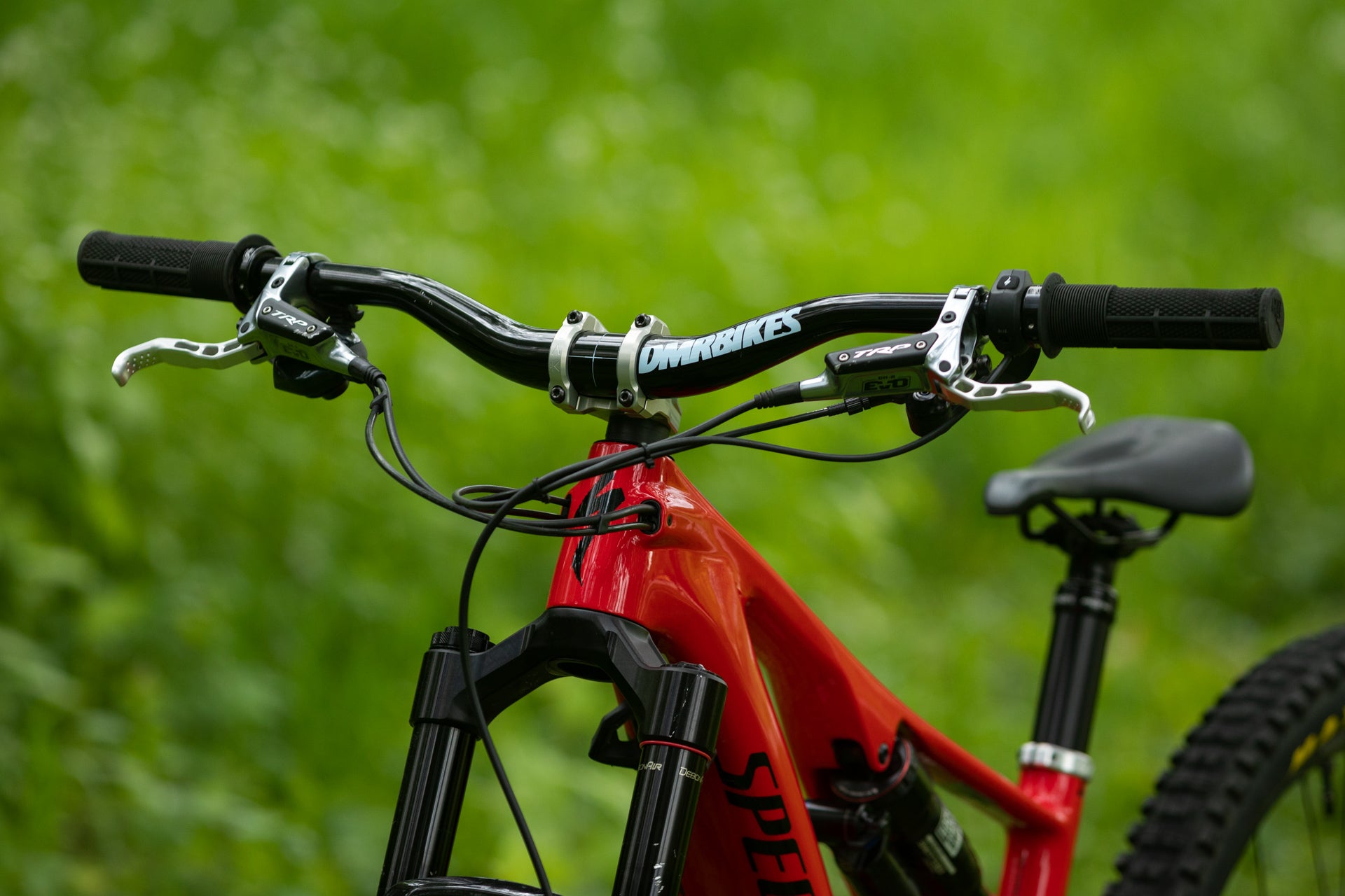 Red mountain bike frame with black handlebars labeled DMRBIKES, partially visible SPE lettering, green foliage