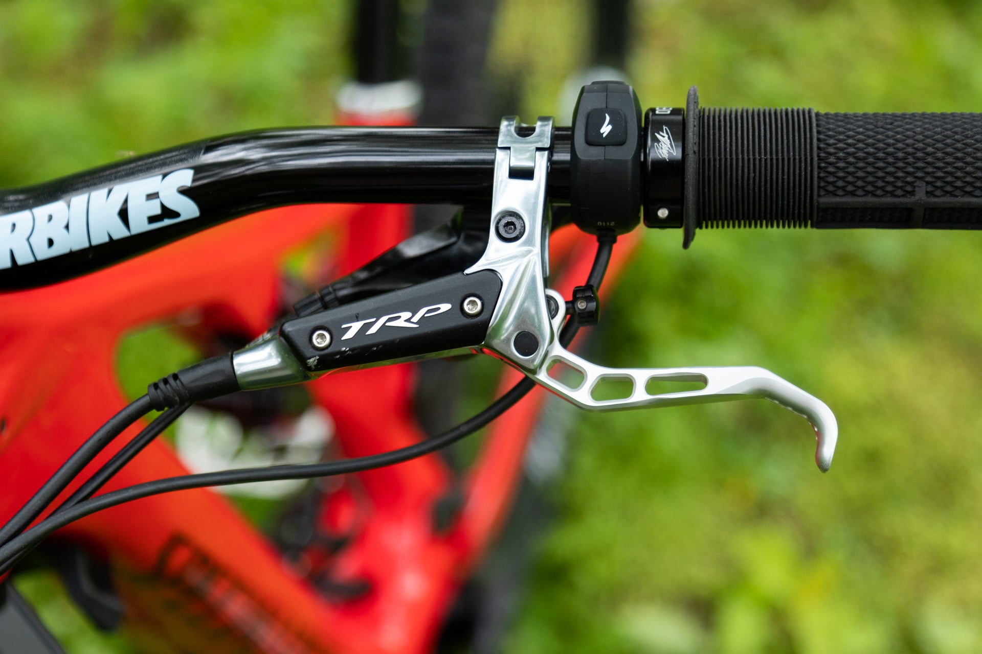 Close-up of TRP brake lever and control button on black handlebar grip, with red RBIKES frame outdoors.