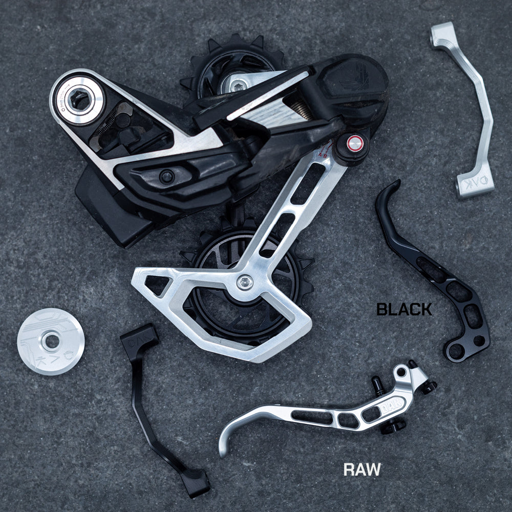 Disassembled mountain bike derailleur, silver 'RAW' brake lever, black 'BLACK' lever, on dark background.