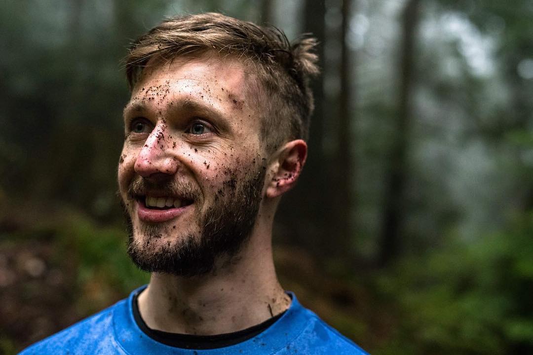 Smiling young man with muddy face and beard in blue shirt, blurred green forest background.