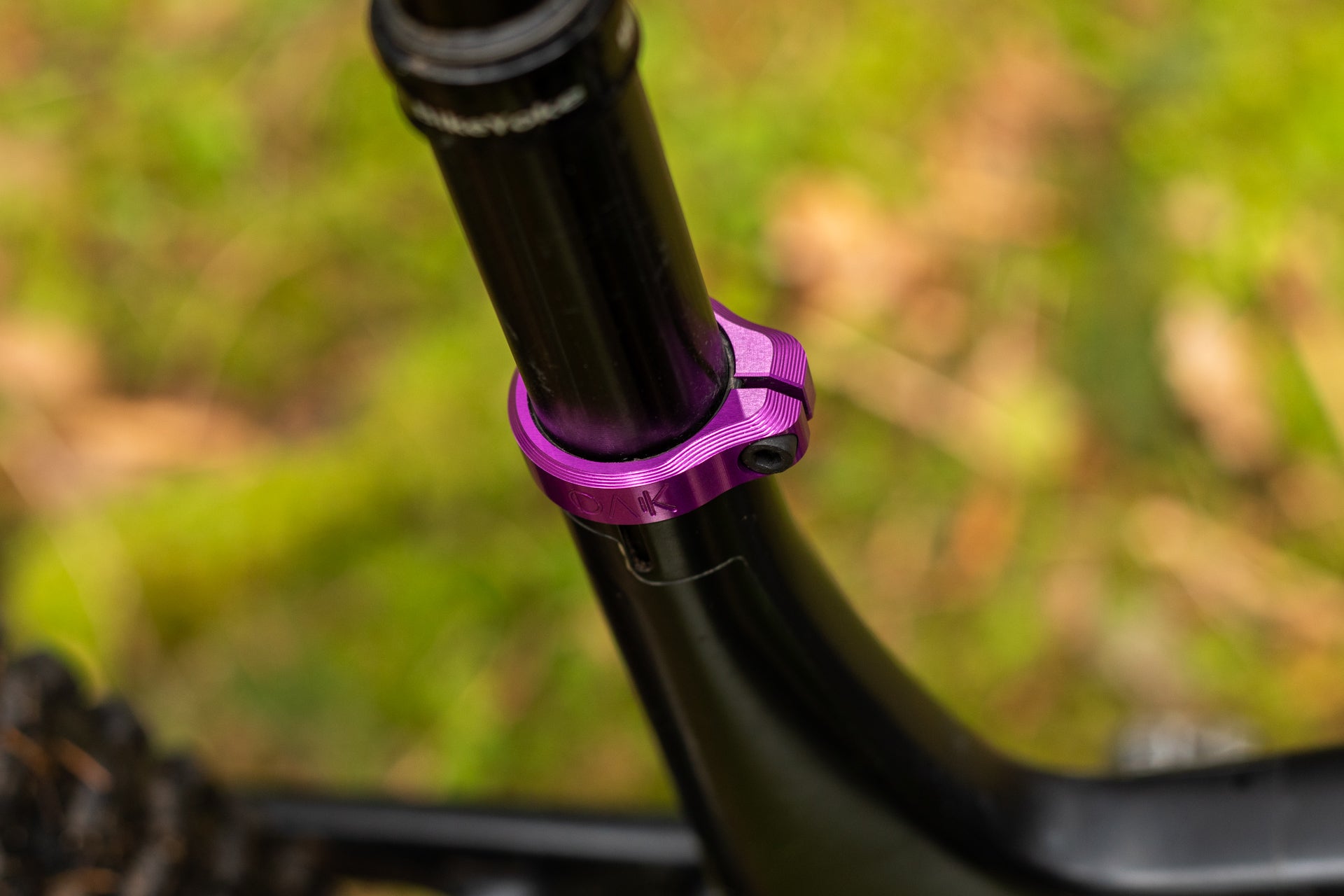 Black bicycle seat post with a purple metallic OAK clamp on a matte black frame outdoors.
