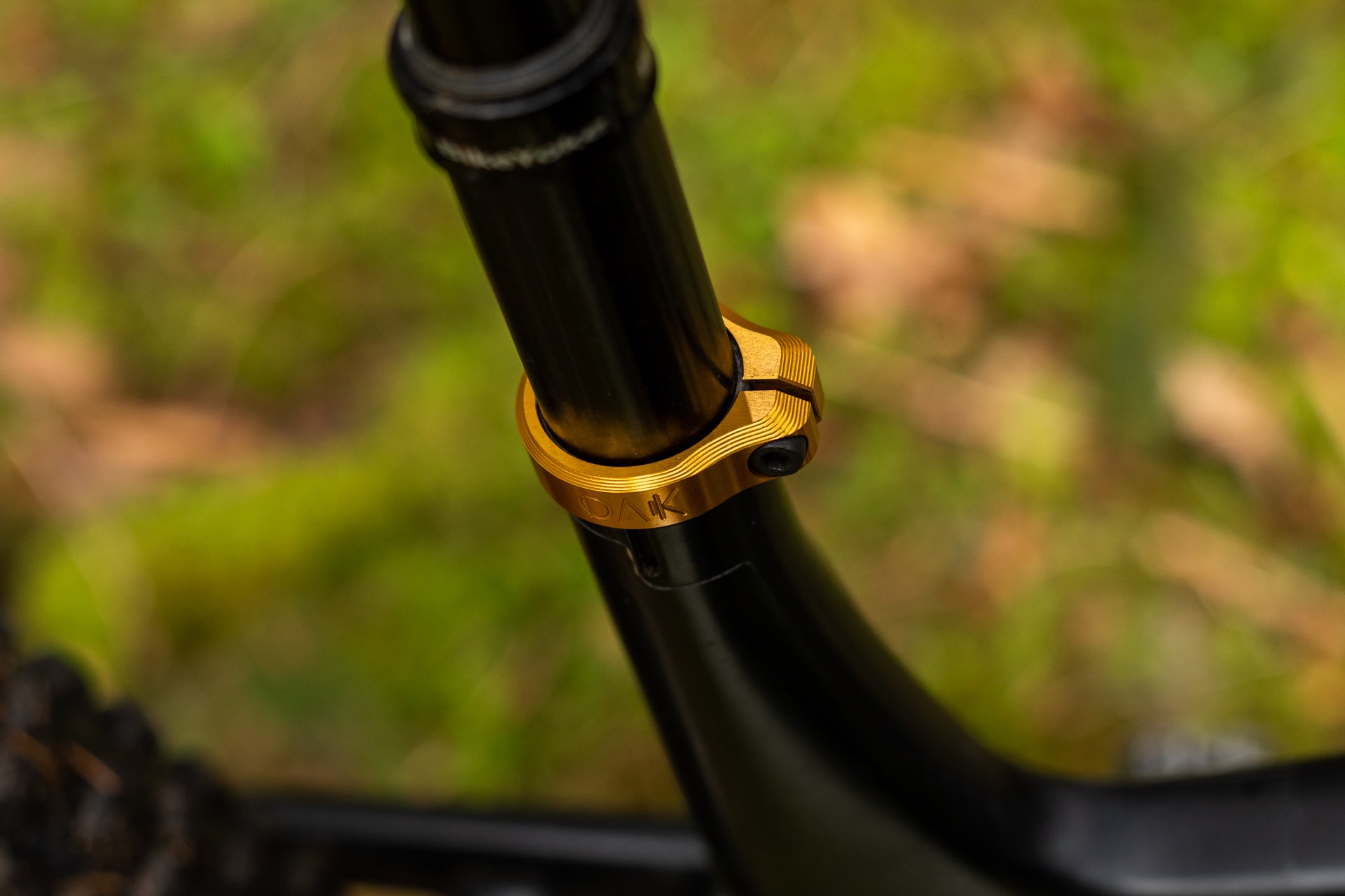Black bicycle seat post with gold OAK engraved clamp against a blurred green background