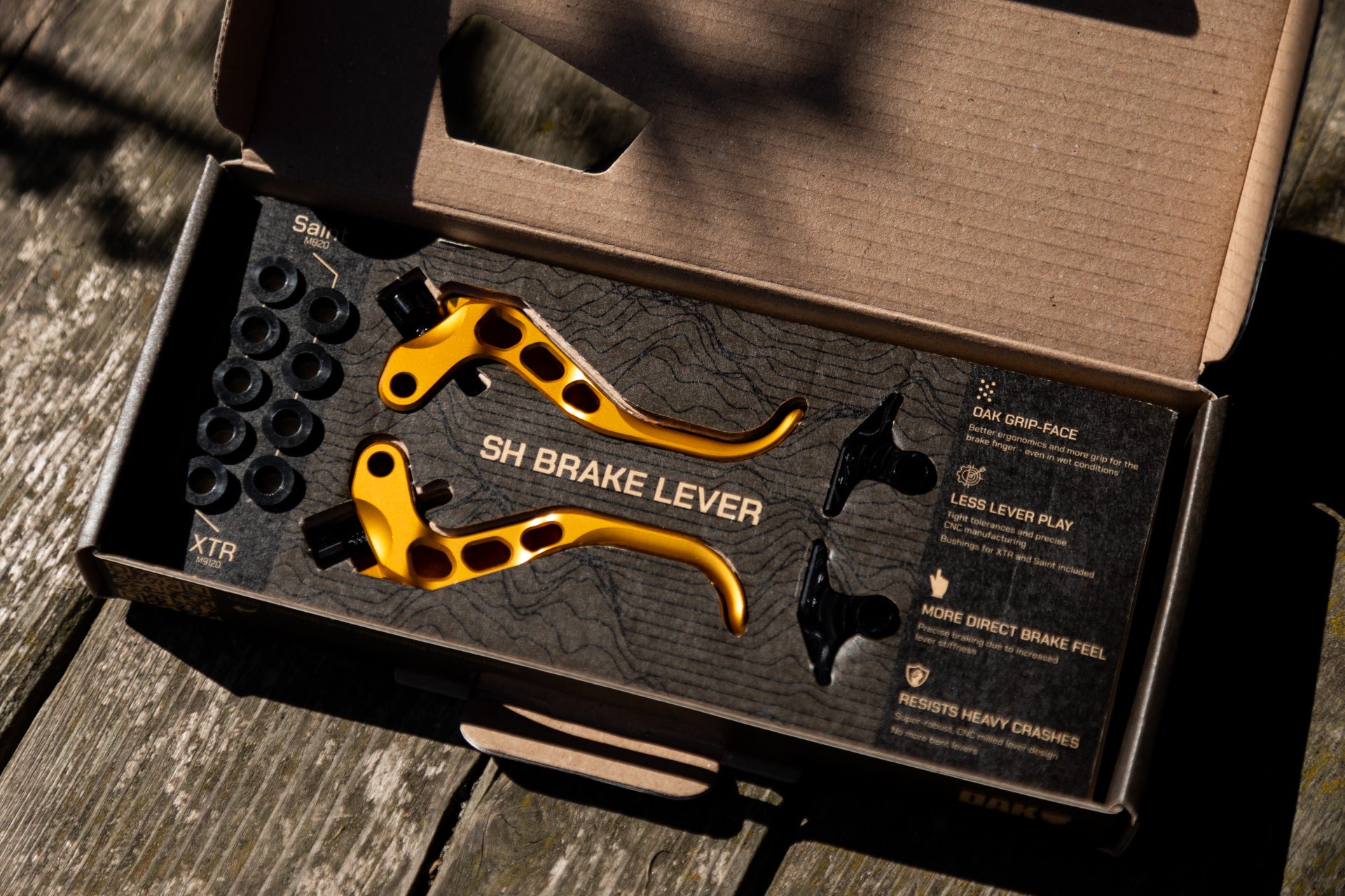 Gold SH brake levers with black bushings labeled M820 and XTR, with black ergonomic parts in box on wood surface.