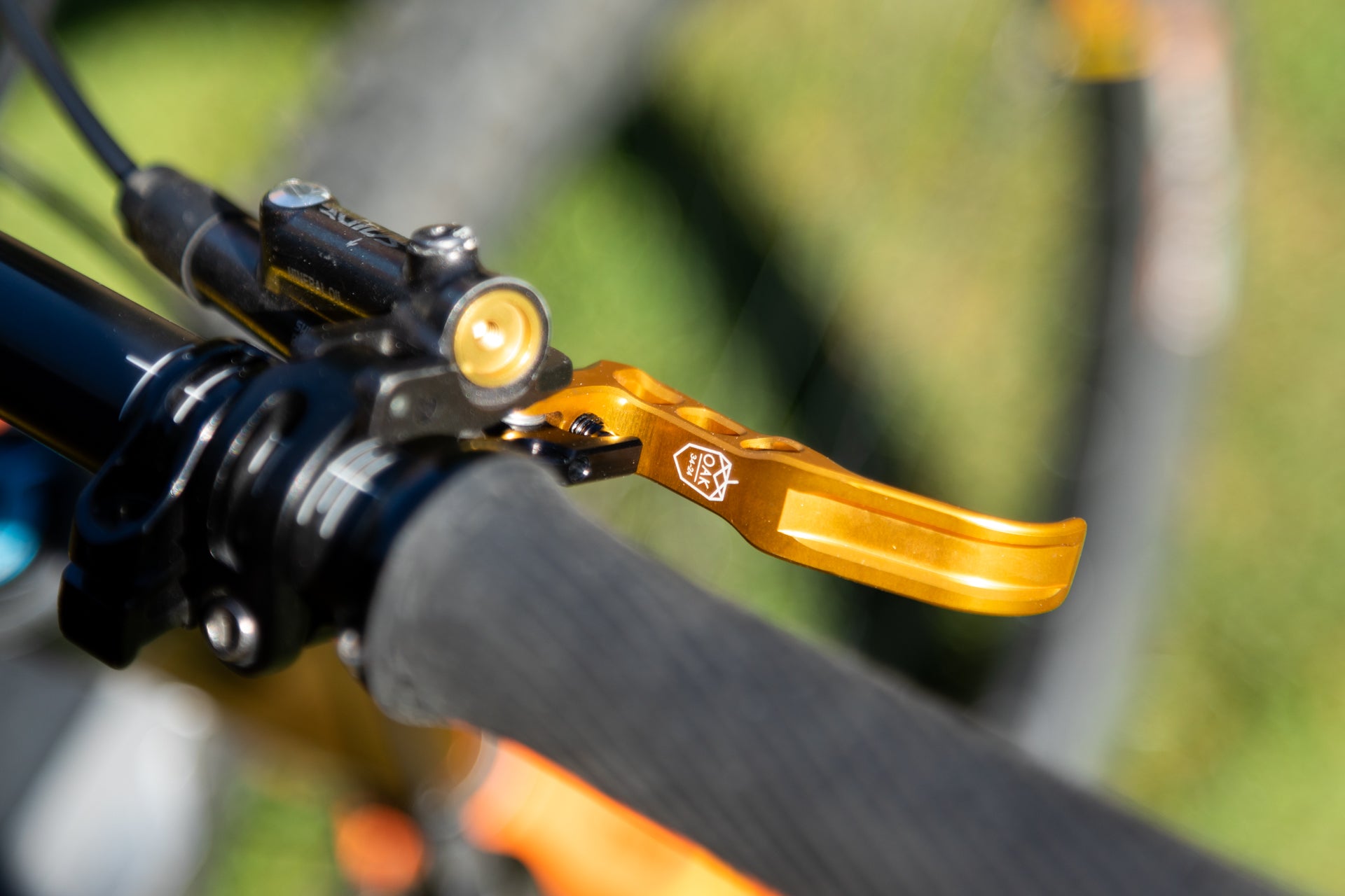 Golden-orange OAK bicycle brake lever, white logo, black textured handlebar grip, black brake assembly, gold adjuster knob.
