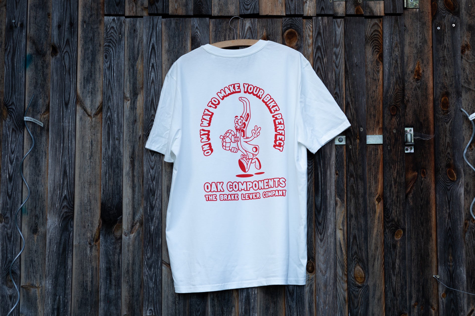 White T-shirt with red OAK COMPONENTS logo and cartoon brake lever holding a wrench on wooden background.