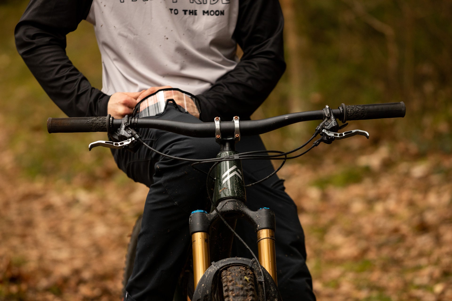 Person in 'TO THE MOON' jersey on a mountain bike with golden suspension forks on a forest trail