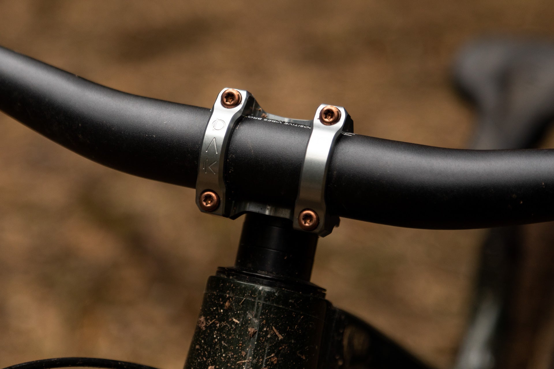 Black mountain bike handlebar with silver clamp and bronze screws, text 'POSITIVE VIBES BRO', dirt-covered frame below.