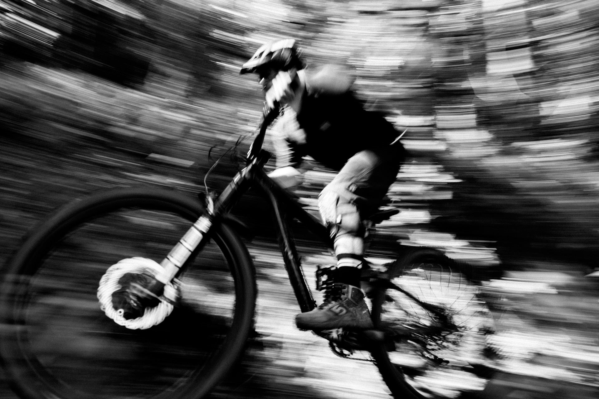 Mountain biker wearing helmet and gear rides swiftly through wooded trail, motion blur on bike and trees.
