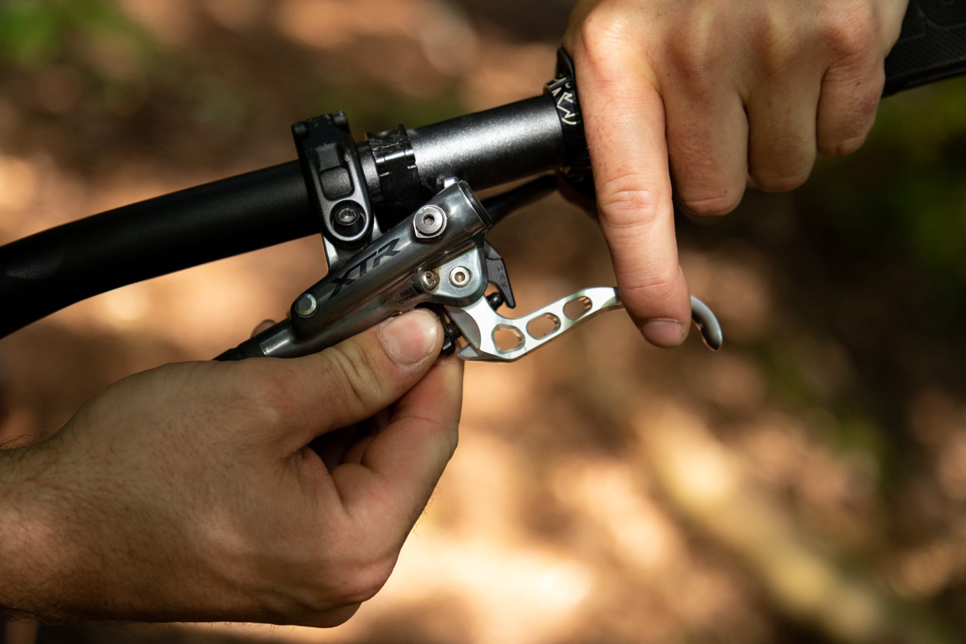 Hands adjusting a silver perforated XTR bicycle brake lever on a black handlebar.