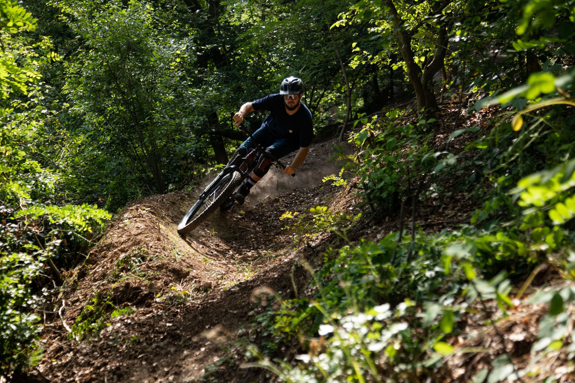 A male mountain biker in a black helmet and sunglasses leaning into a curve on a sunny forest trail.