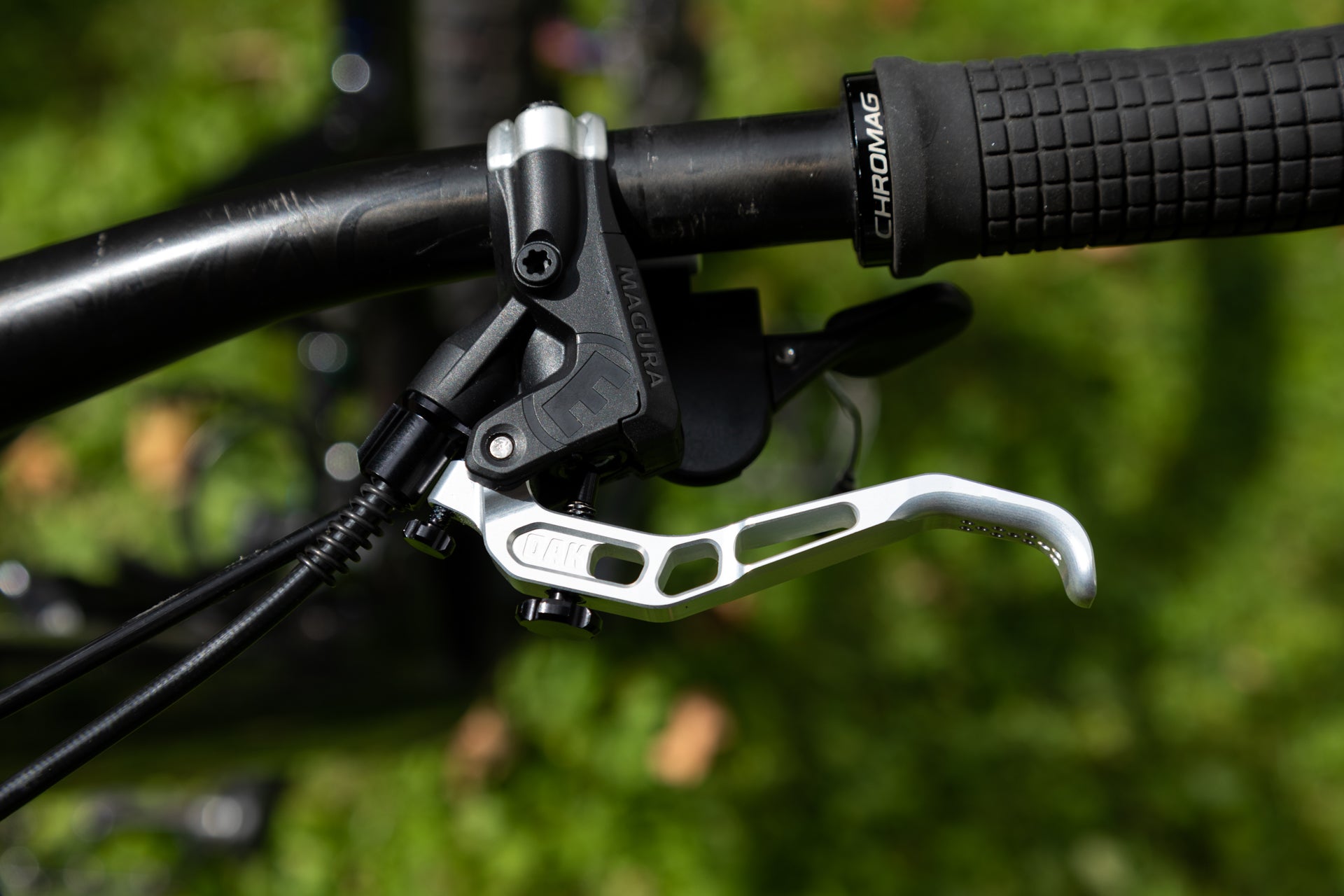 Close-up of a black MAGURA brake lever and metallic lever on a scratched black handlebar with a CHROMAG grip, outdoors.