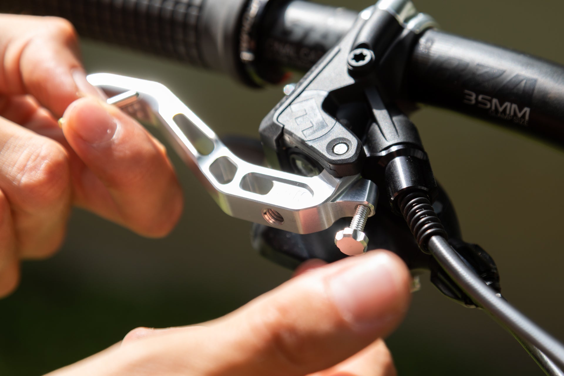 Hand adjusting a shiny metallic brake lever on a black 35mm bicycle handlebar with blurred green background.