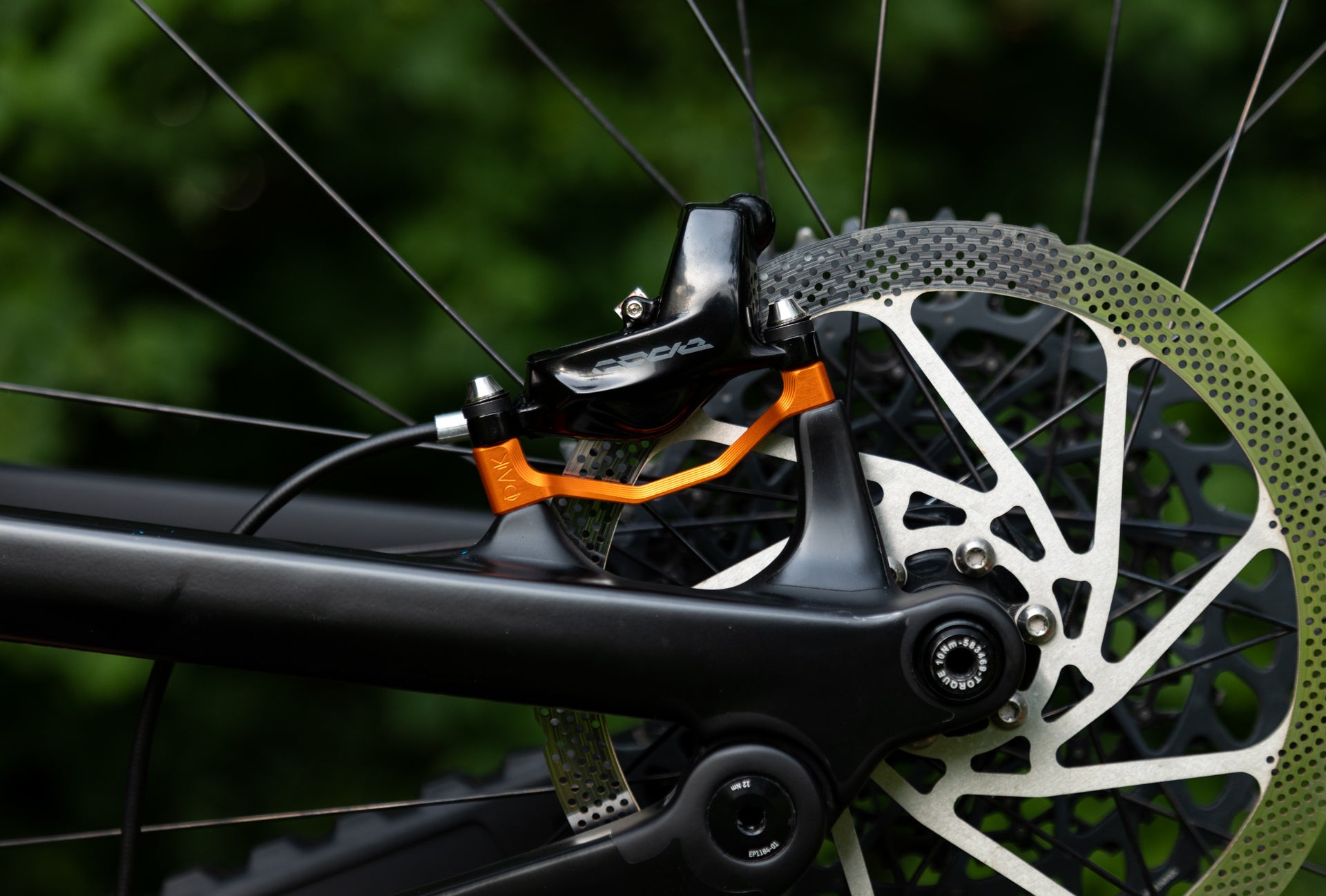 Close-up of a black rear disc brake caliper with an orange adapter on a matte black bike frame and silver rotor.