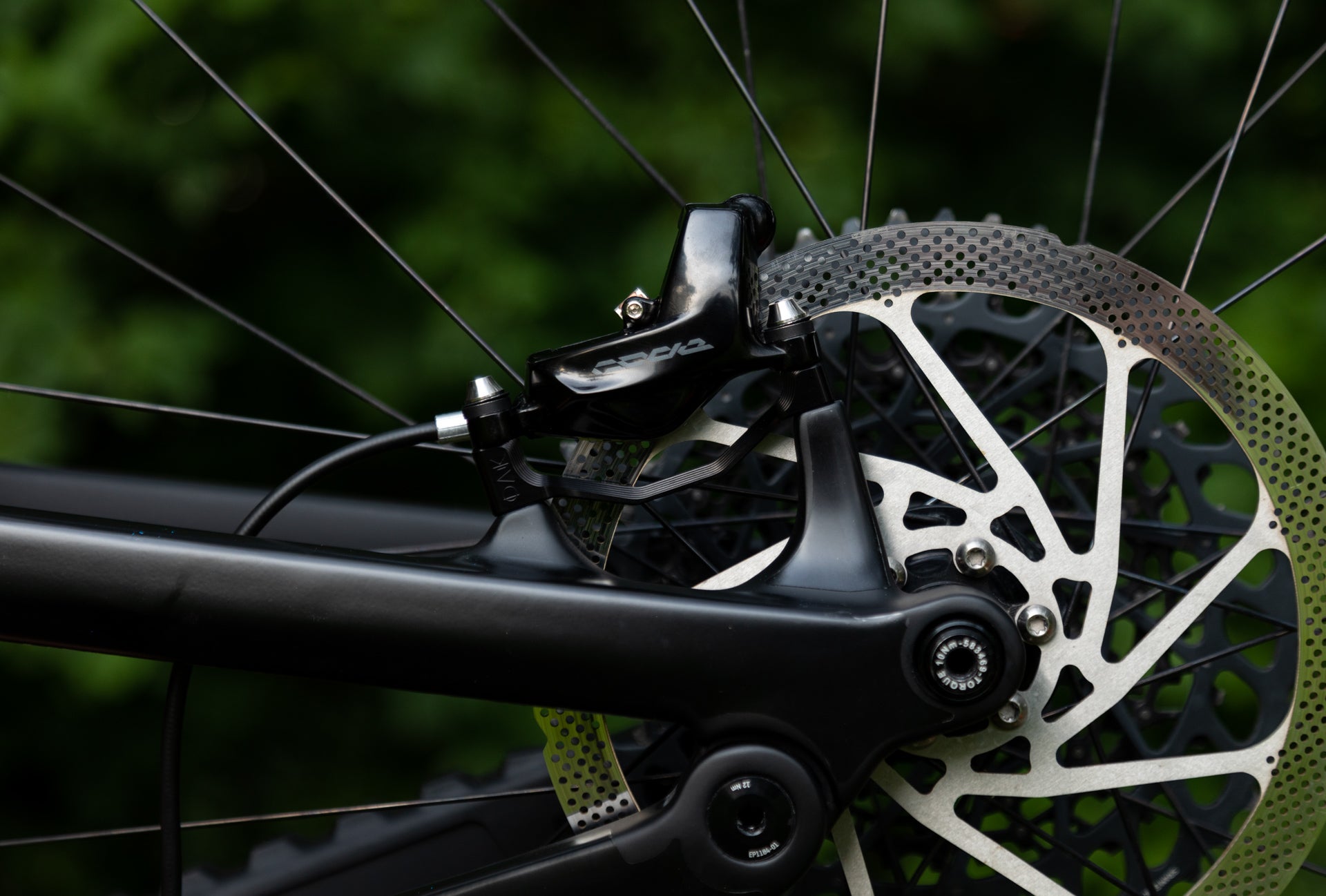 Close-up of matte black rear bicycle disc brake with caliper and silver rotor, blurred green foliage behind.