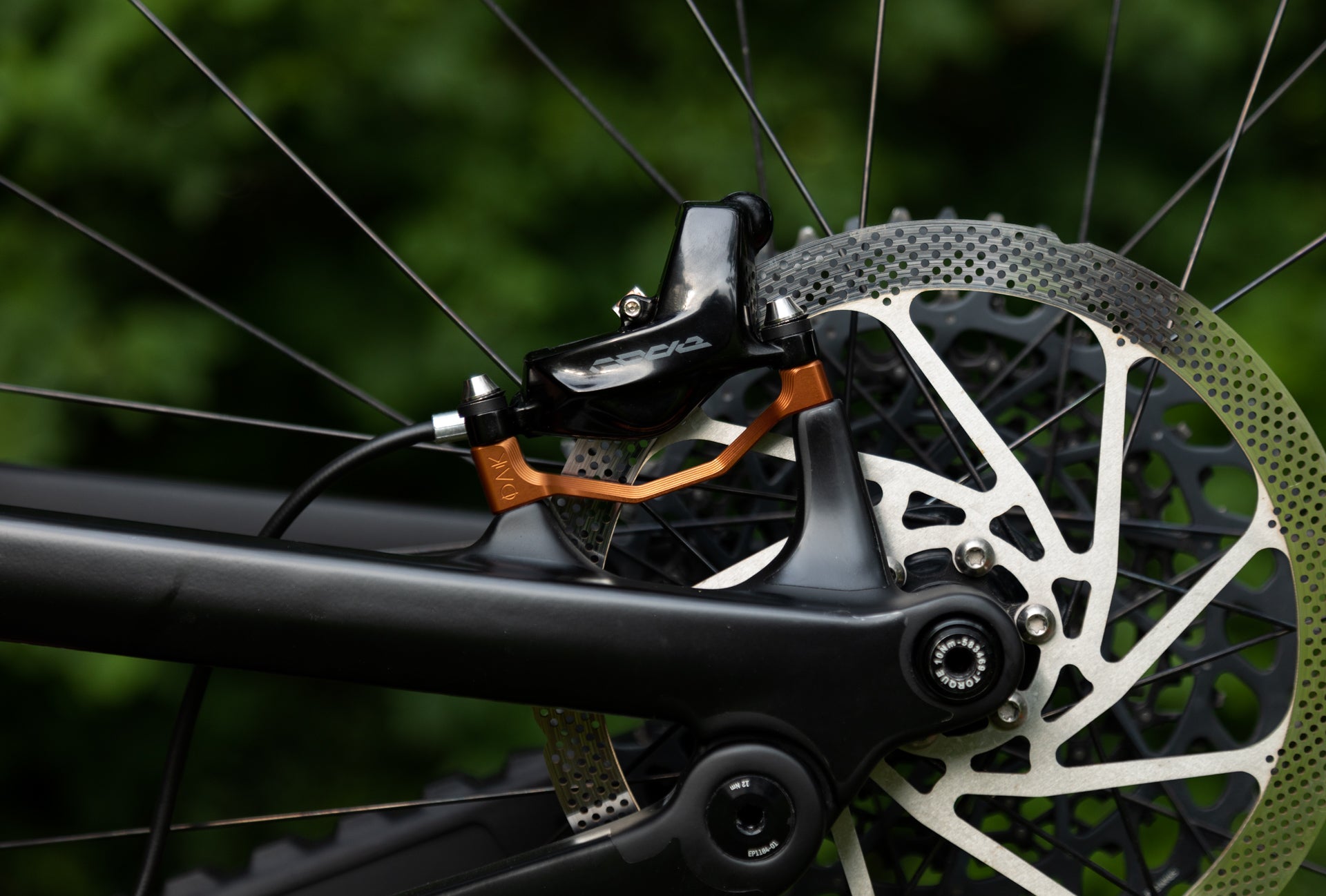 Close-up of a black rear bicycle disc brake caliper with orange hydraulic adapter on silver perforated rotor.