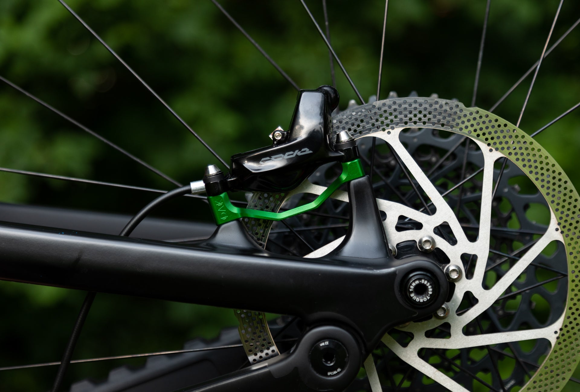Close-up of bike disc brake with black caliper EP1184-01, green arm, and perforated rotor on matte black frame.
