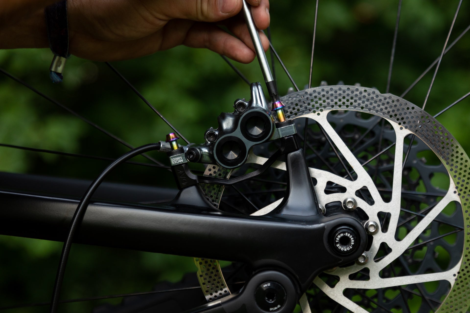 Hand adjusting black rear disc brake caliper on mountain bike with matte frame and silver rotor marked CAN