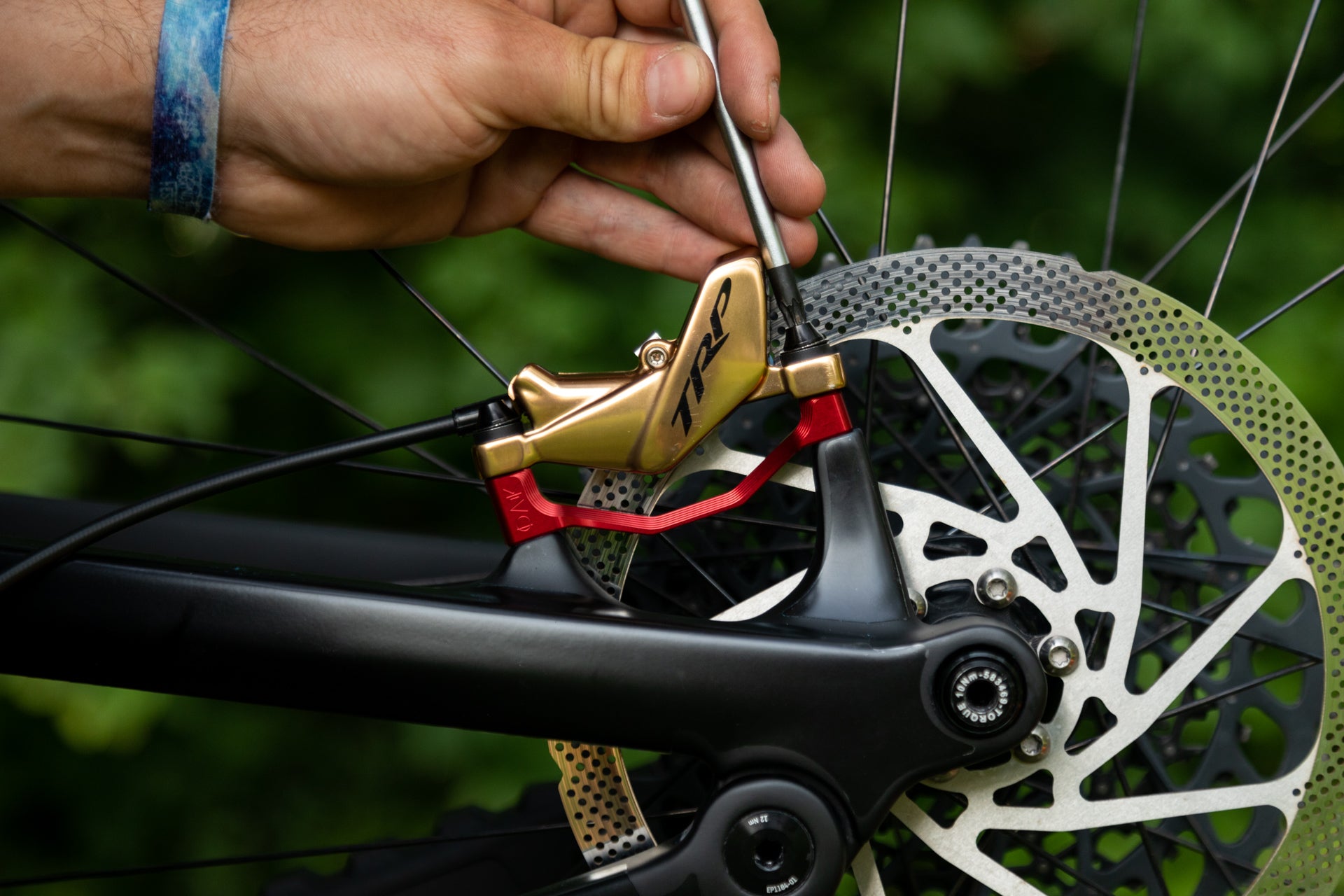 Metallic gold TRP bike disc brake caliper with red lever on black frame, hand adjusting with screwdriver.