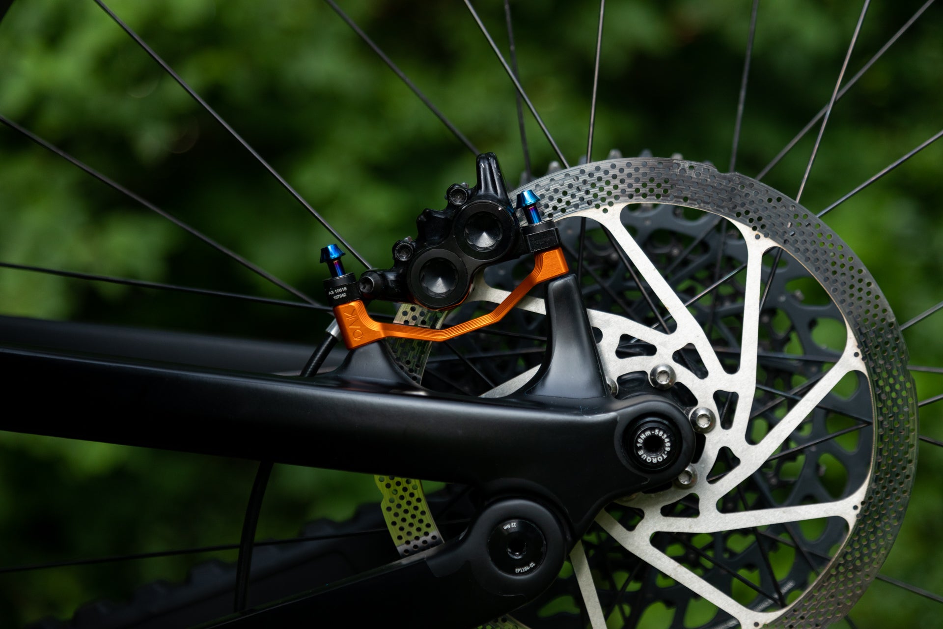 Close-up of a mountain bike rear disc brake with black and orange caliper and silver rotor against green.