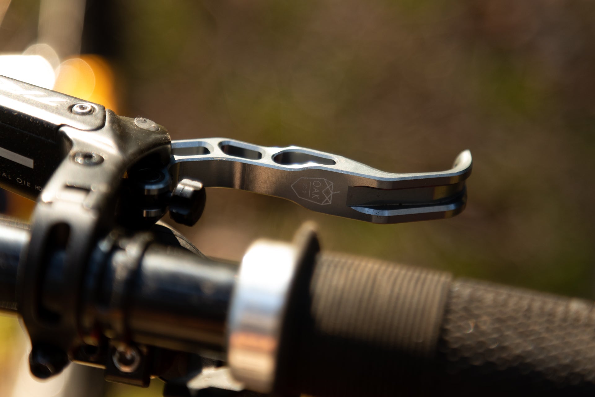 Metallic bicycle handlebar lever with 'OAK' engraving and geometric logo on a textured black grip.