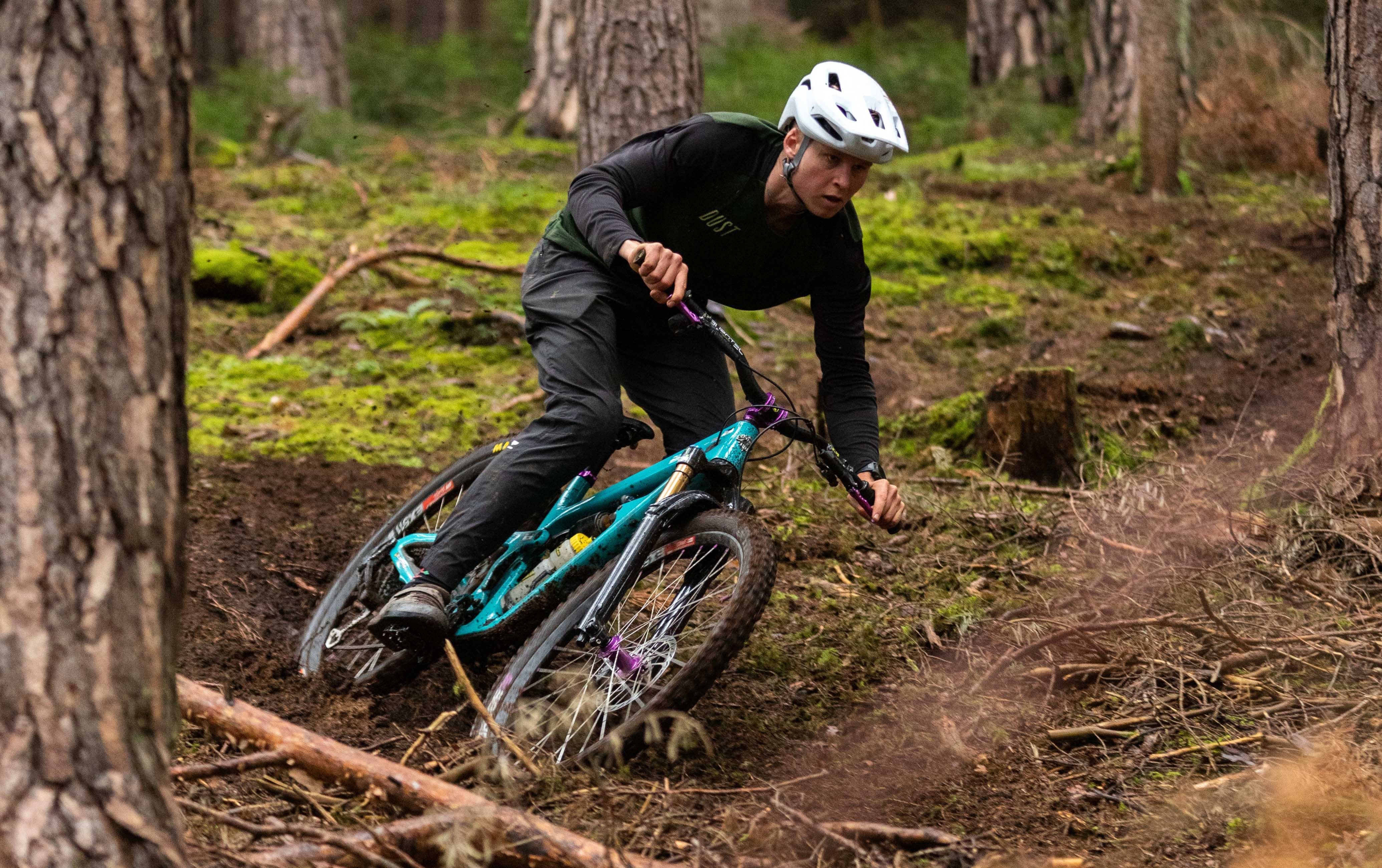 Male cyclist in white helmet and black DUST shirt riding a turquoise mountain bike on a forest dirt trail