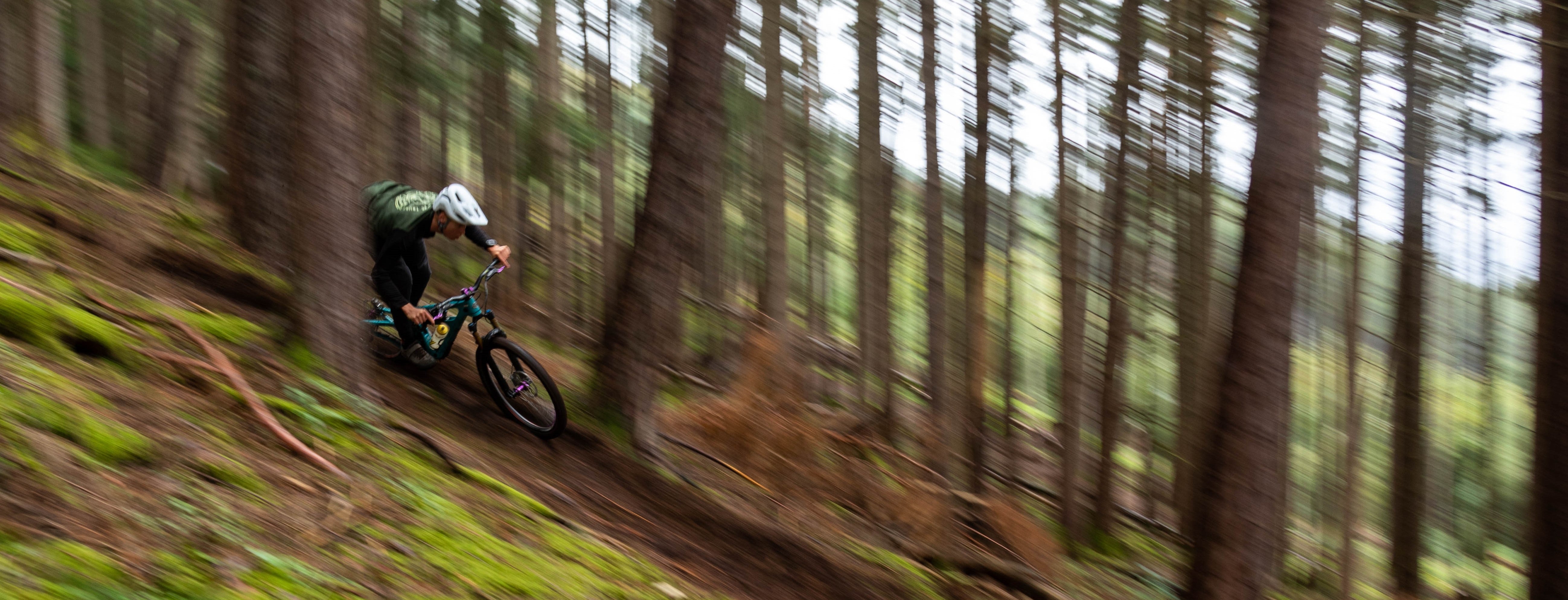 Mountain biker in a white helmet and green jacket rides a blue bike downhill on a forest trail.