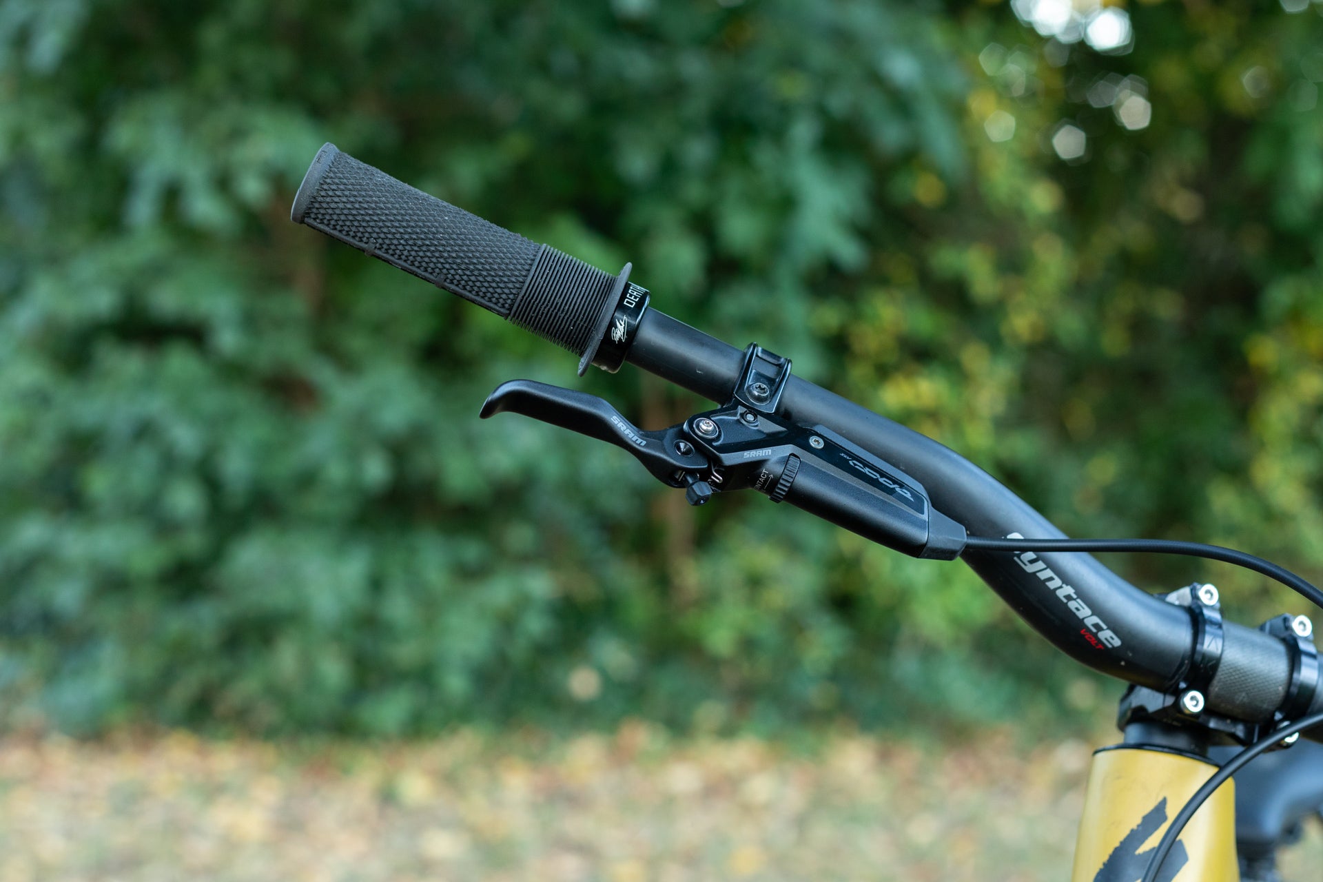 Close-up of a mountain bike's left handlebar featuring DEATH grip, SRAM brake lever, yntace VOLT handlebar on a yellow frame in a natural setting.