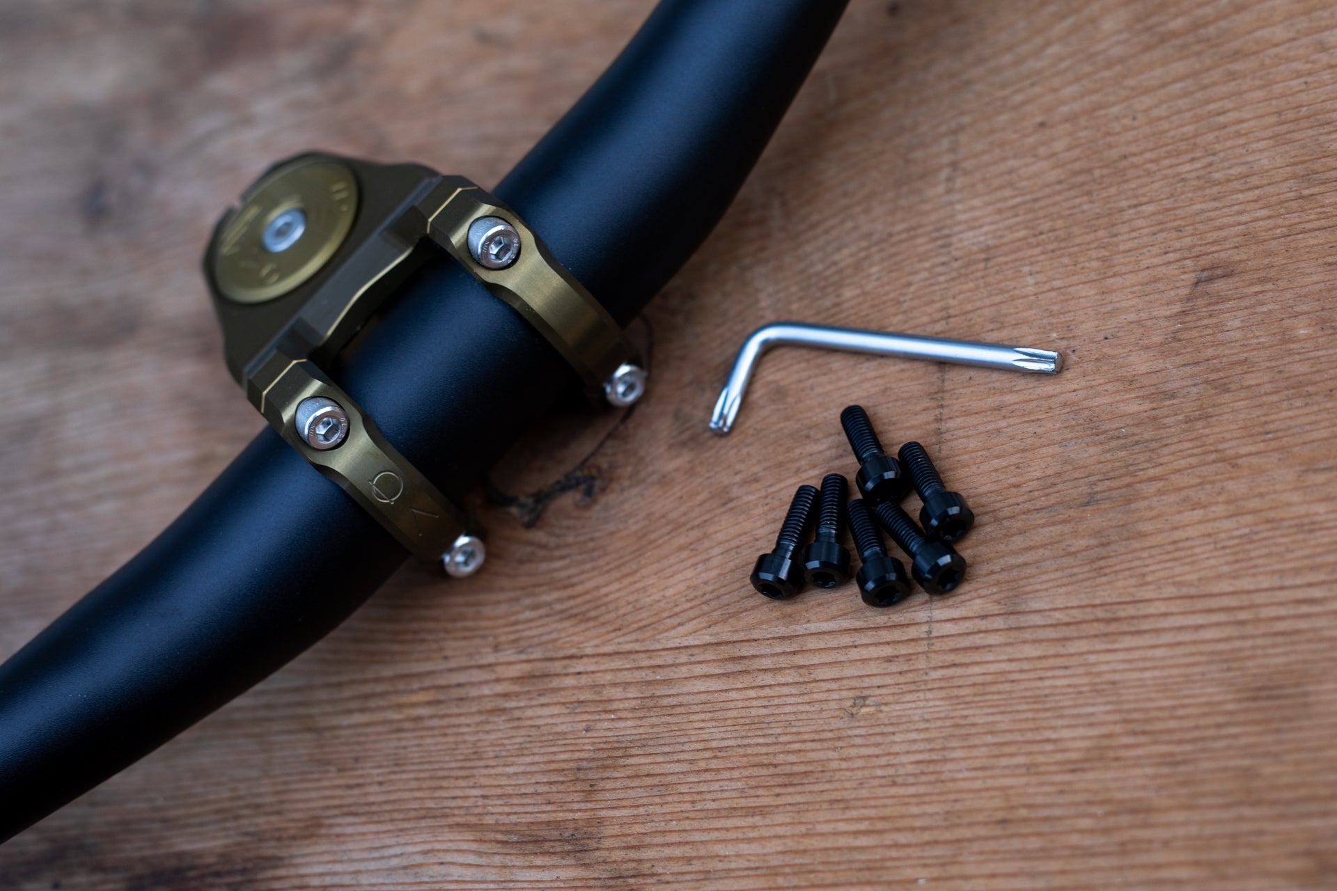 Black mountain bike handlebar with metallic gold stem clamp, silver screws, and Allen key on wooden surface