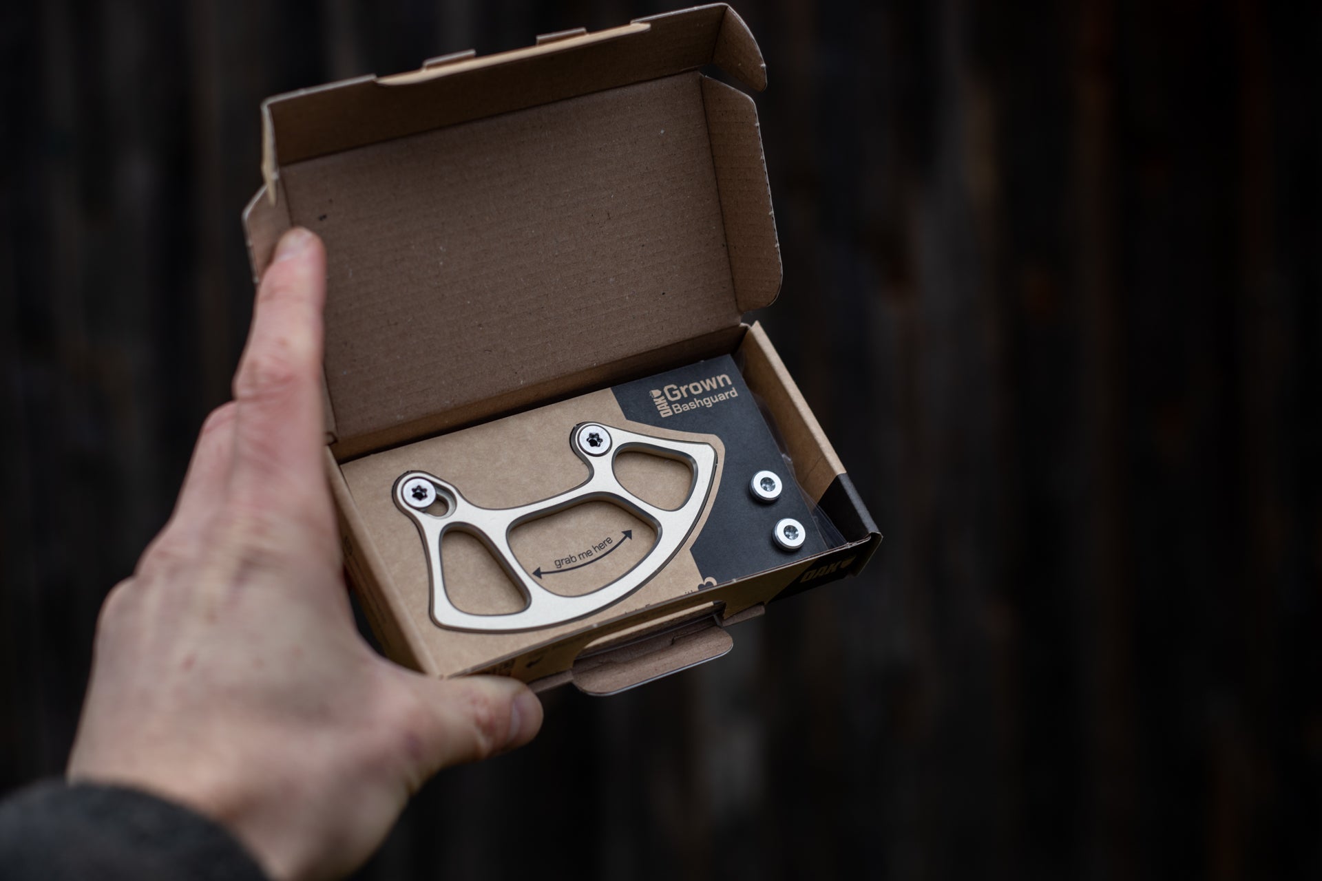 Left hand holding an open cardboard box with a metallic Grown Bashguard and mounting bolts against a dark backdrop