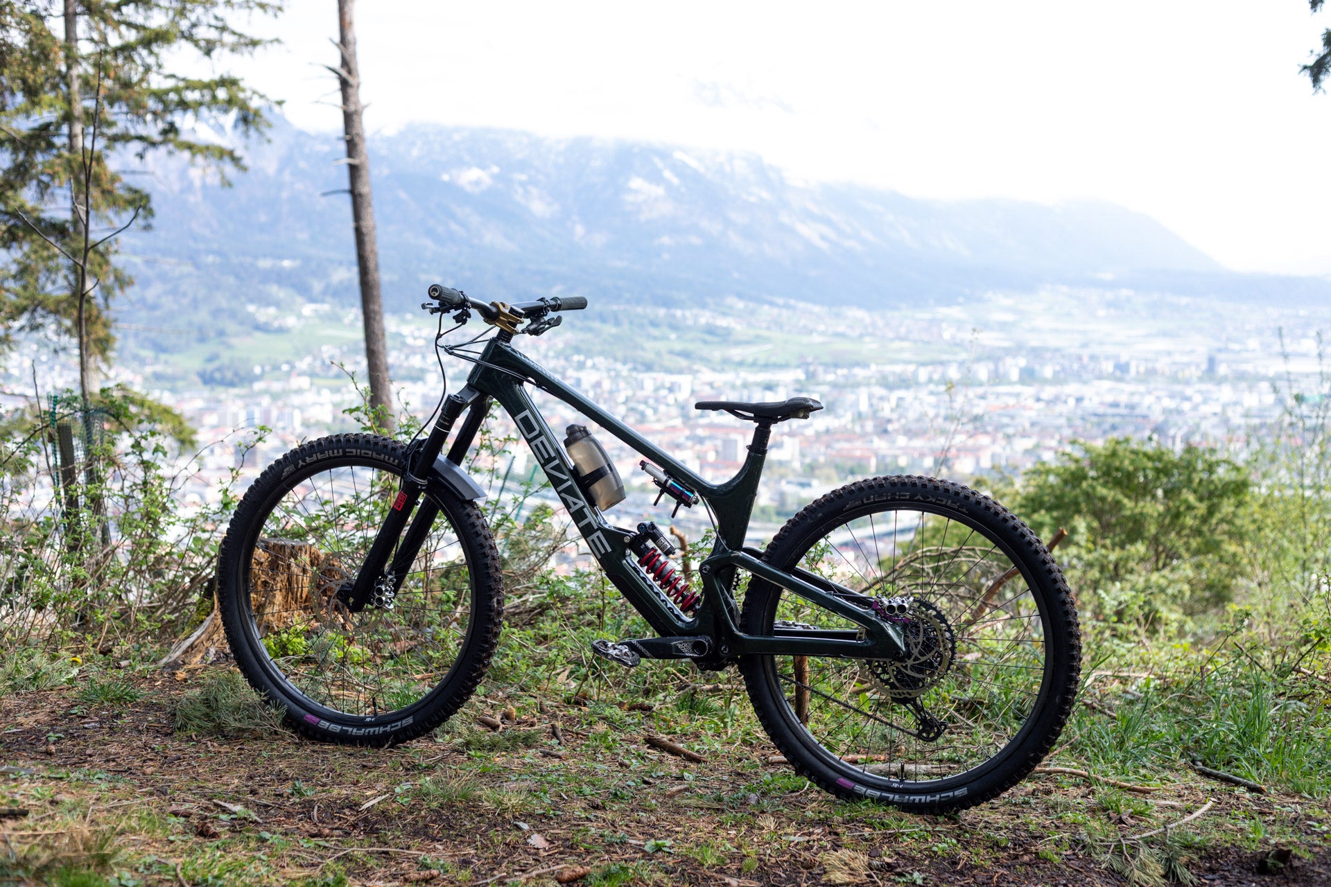 Black full-suspension mountain bike with DEVIATE frame and MAGIC MARY tires on trail near city view.