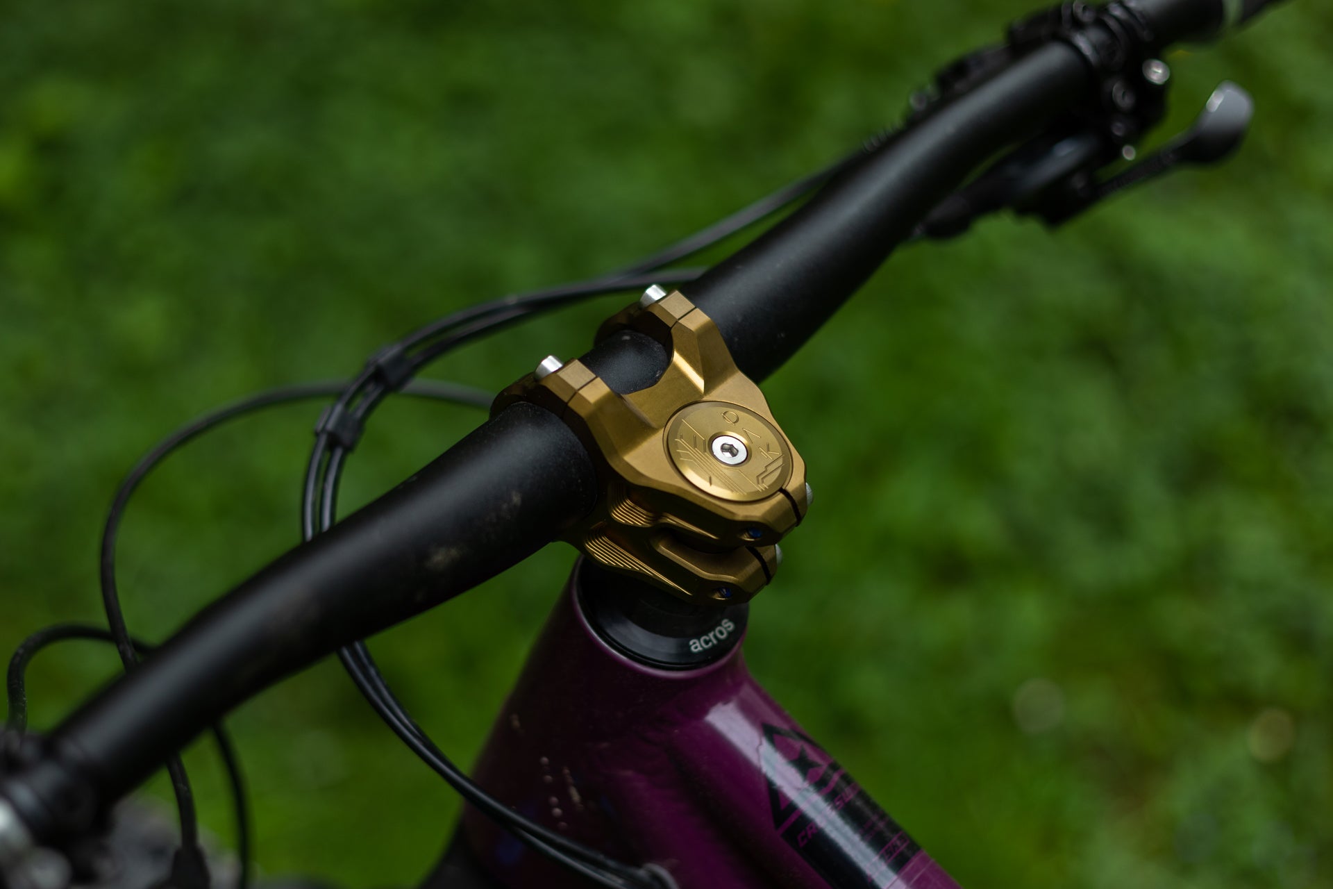 Close-up of a matte black handlebar on a purple mountain bike frame with a gold stem clamp, against a green background.