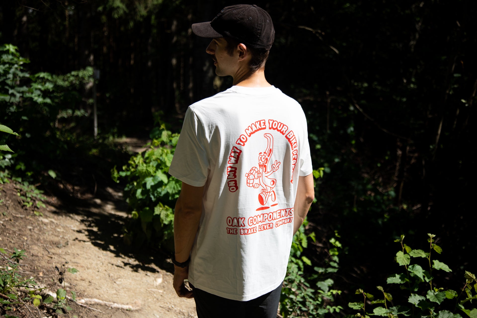 Man on a forest path wearing a white OAK Components T-shirt with red text and a black cap, amidst lush green trees.