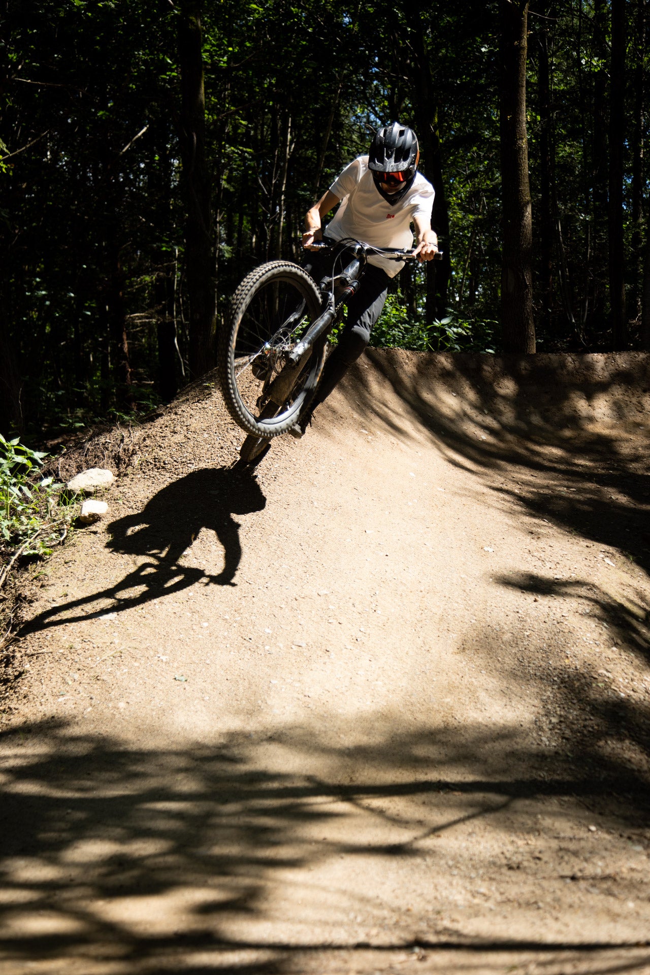 Mountain biker in black helmet and white shirt doing a wheelie on a forest dirt trail berm in sunlight
