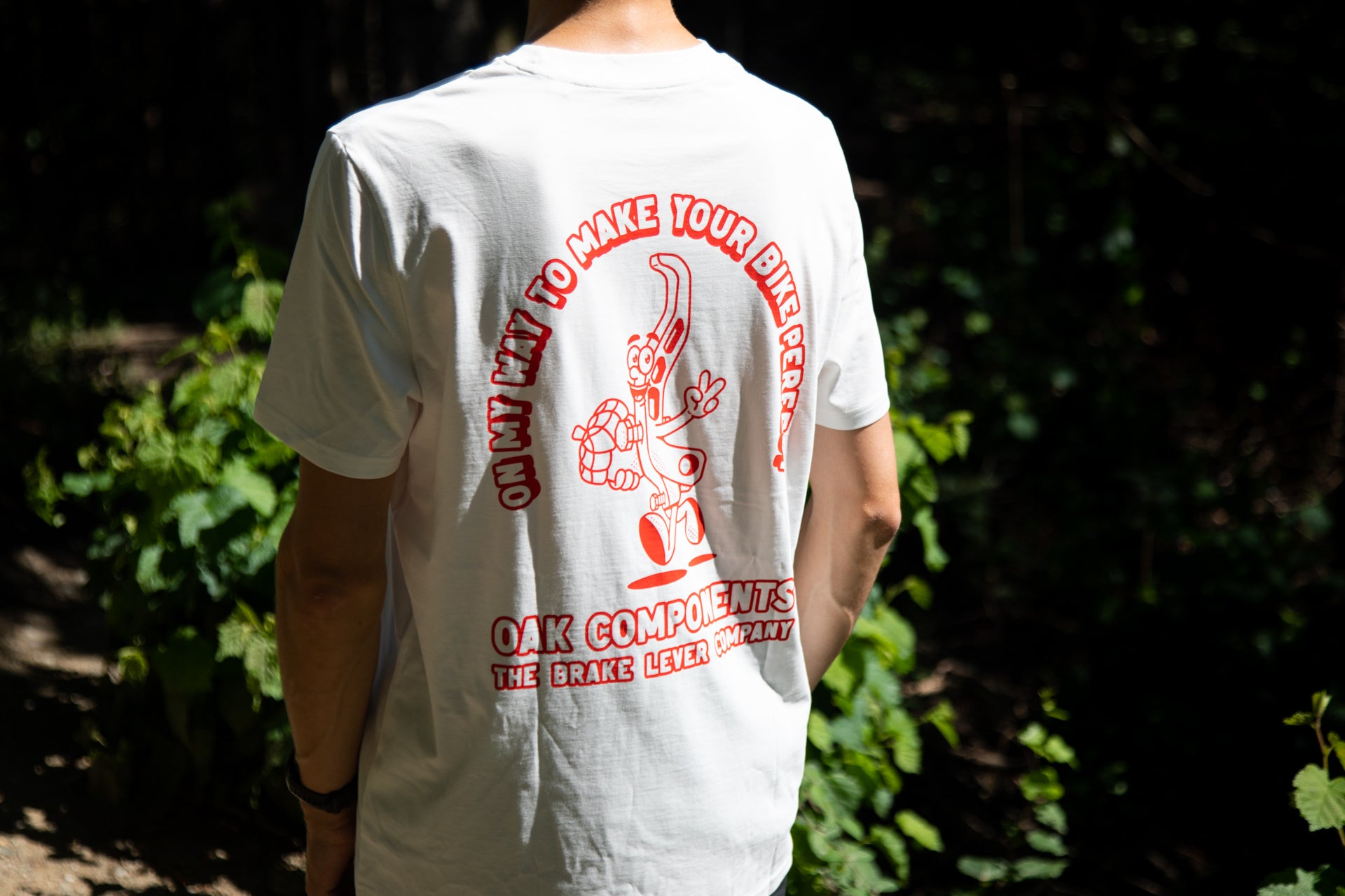 Person outdoors wearing white T-shirt with red cartoon brake lever and 'OAK COMPONENTS' text, green foliage background