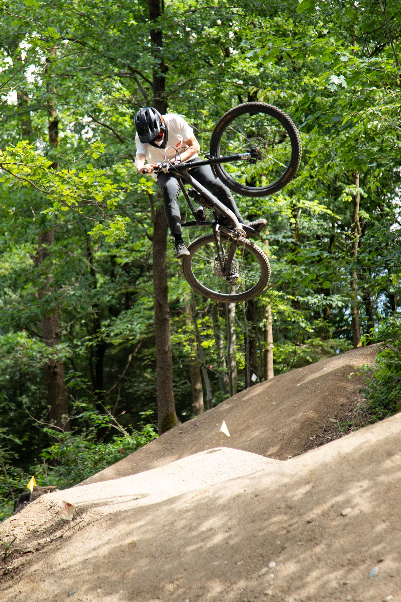 Mountain biker in a black helmet and white shirt jumping over a dirt trail in a forest.