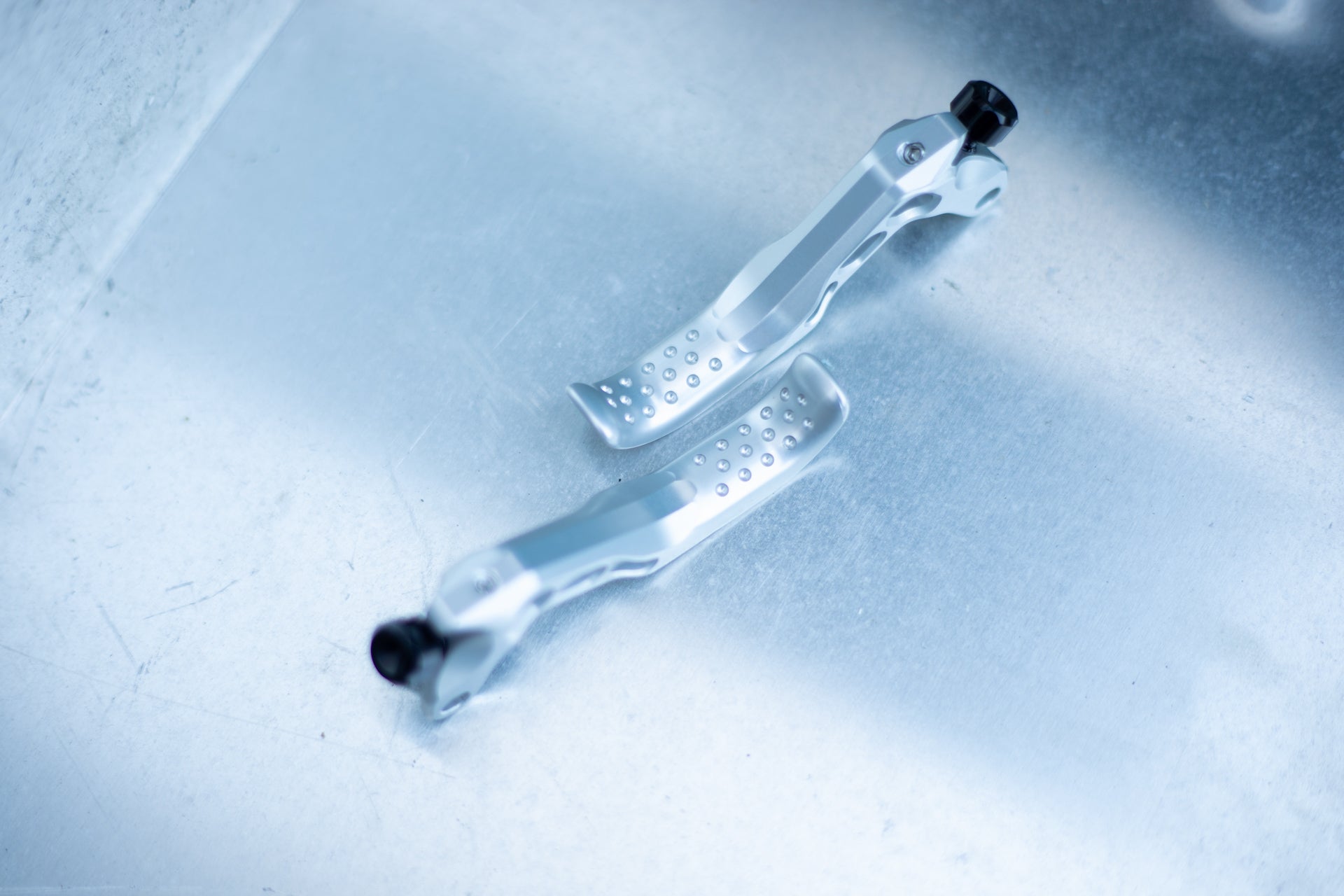 Silver metallic bike brake levers with textured grips and black knobs on a light blue surface.