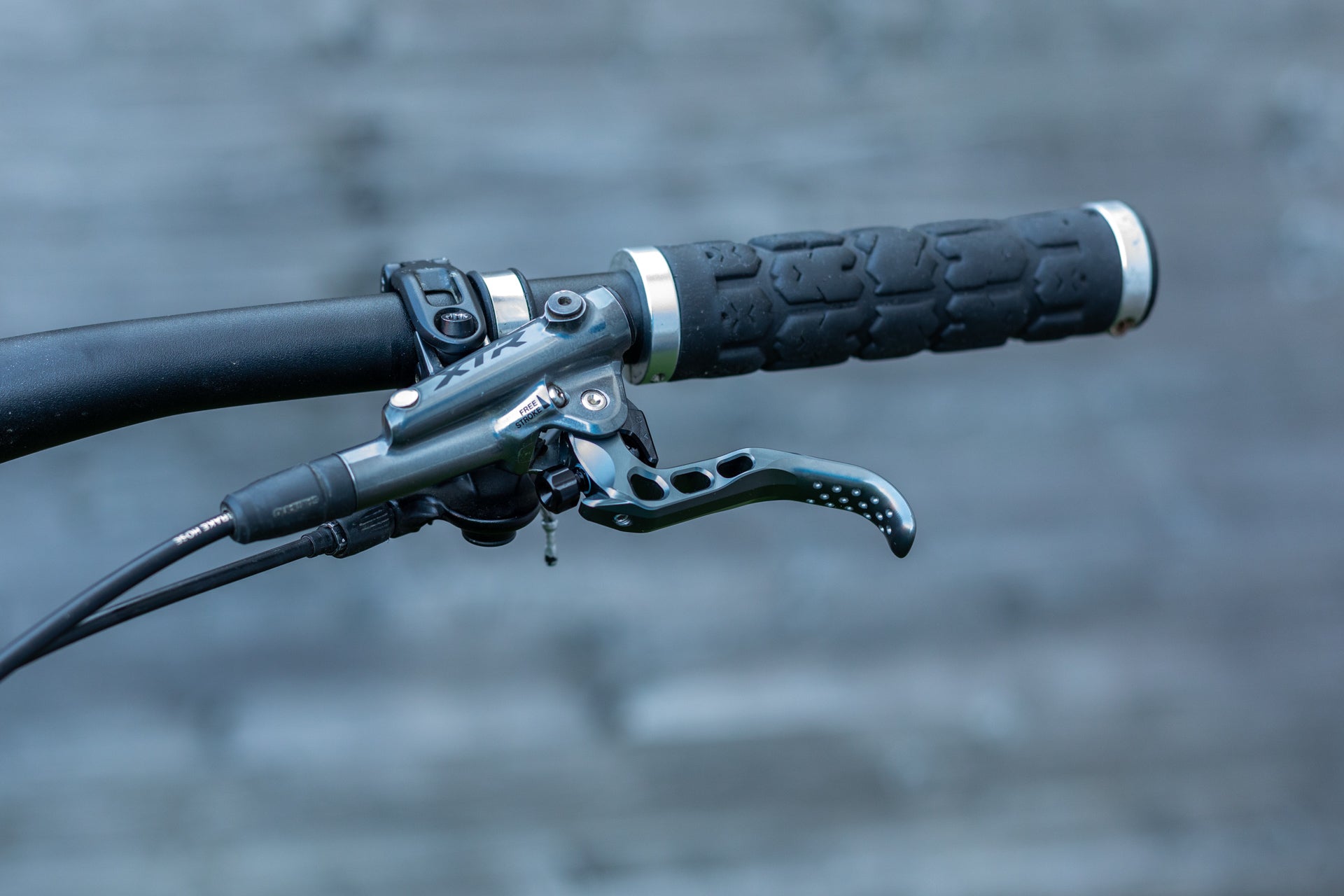 Close-up of mountain bike handlebar with textured rubber grip, curved metallic brake lever, and brake hose.