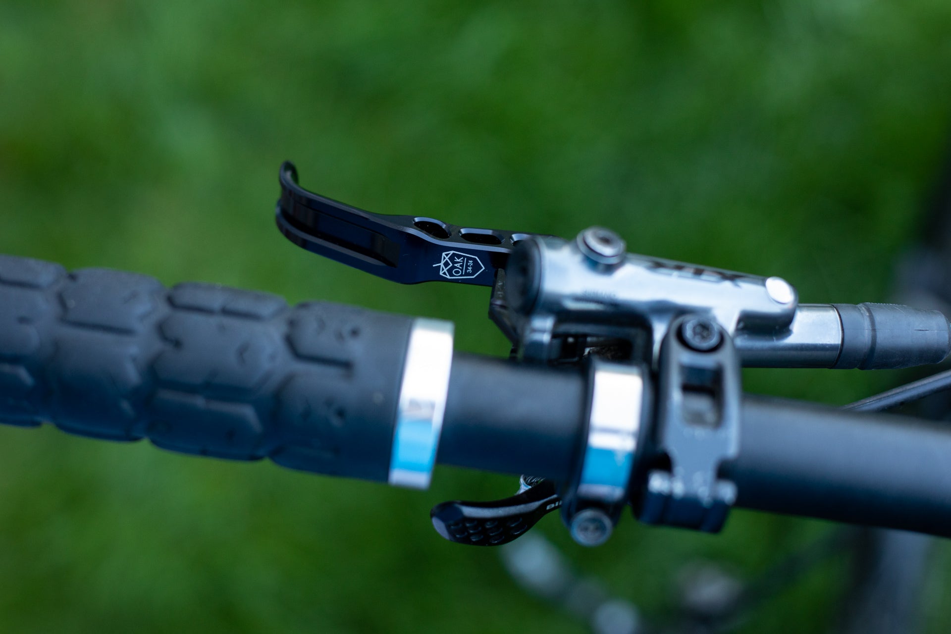 Close-up of mountain bike handlebar with black rubber grip, silver clamp, and OAK brake lever on grass