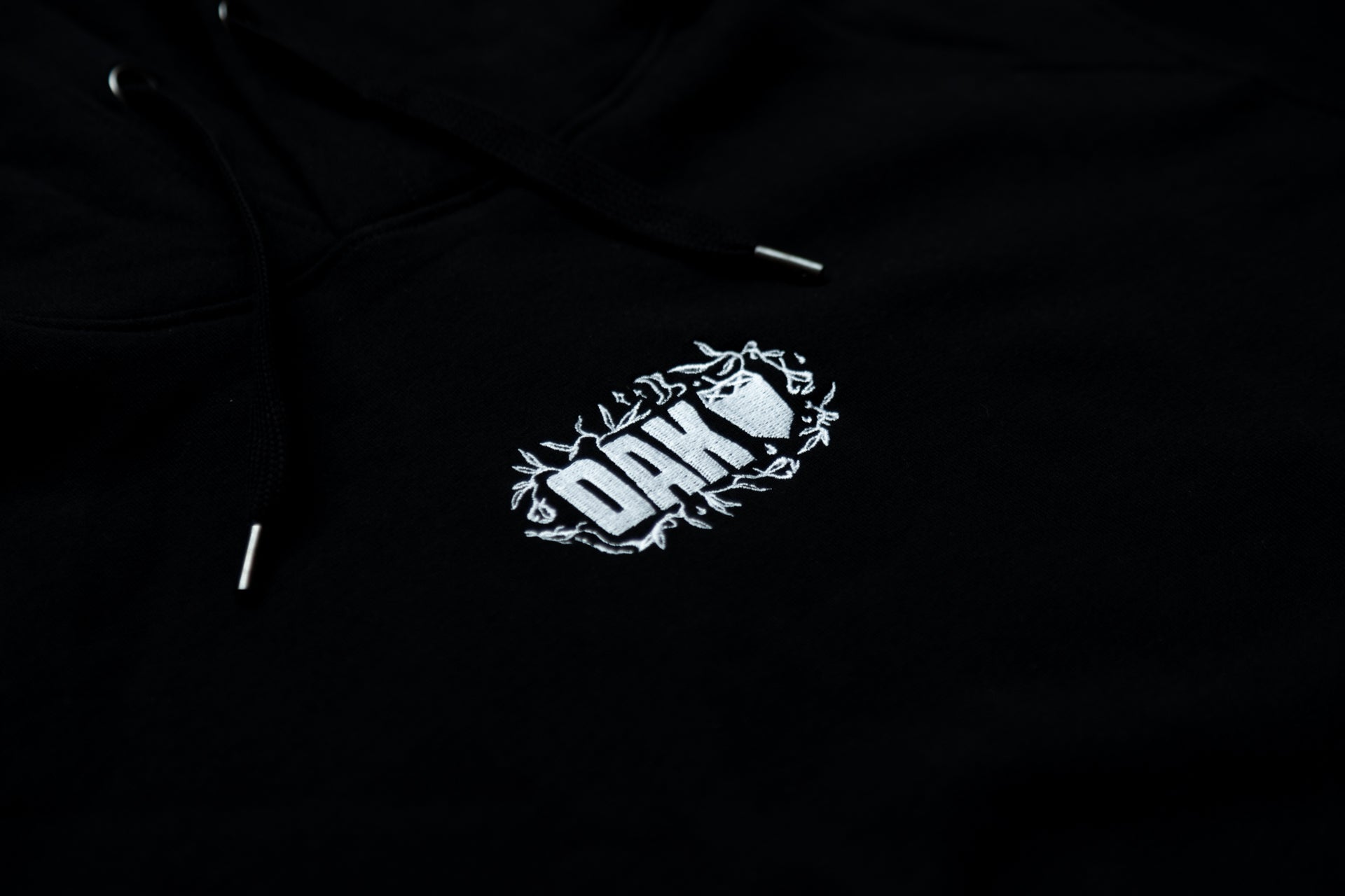 Black hoodie with white embroidered 'OAK' text and leaf pattern on chest, black drawstrings.