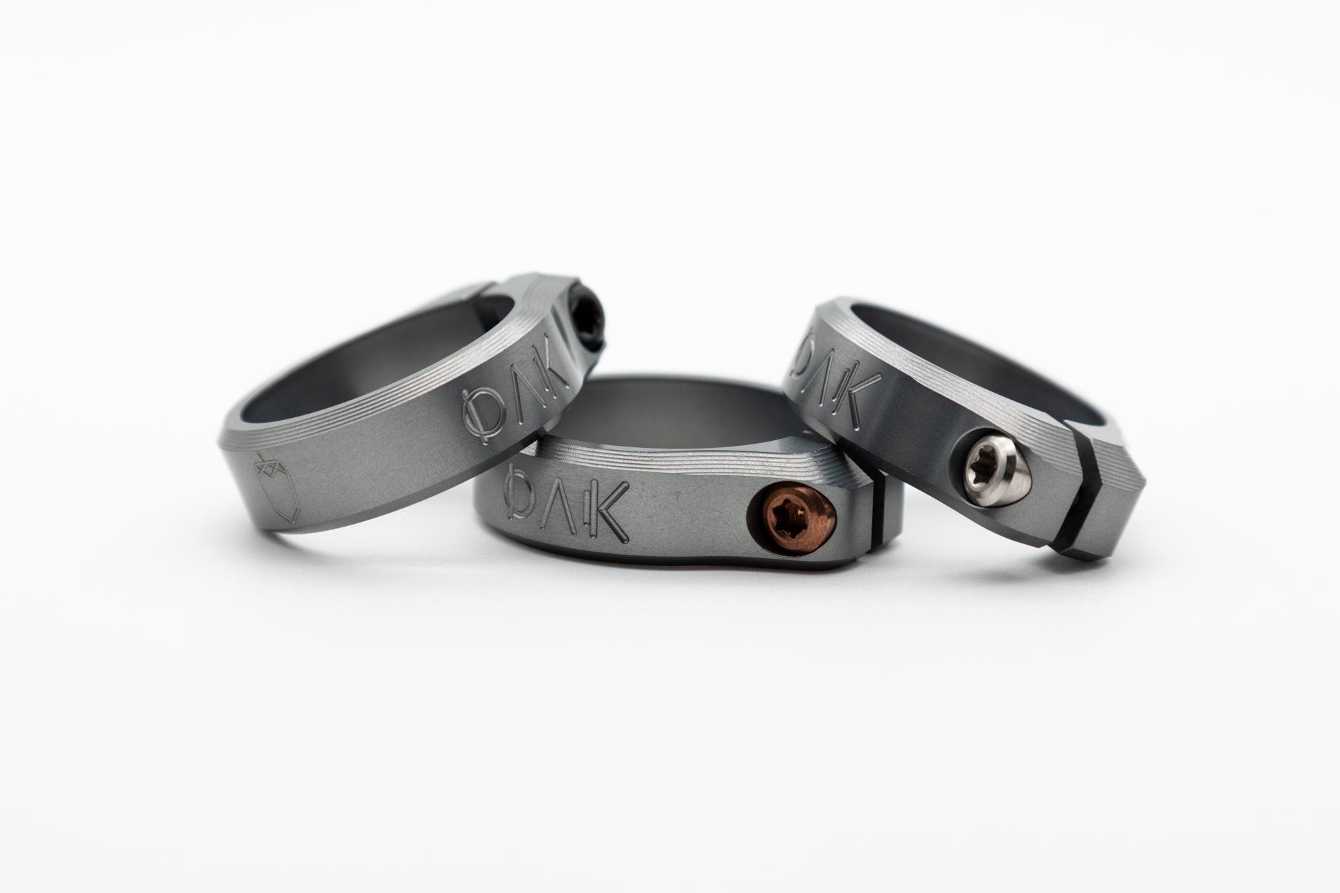 Three metallic mountain bike clamps with engraved OAK branding, featuring copper, silver, and black screws.