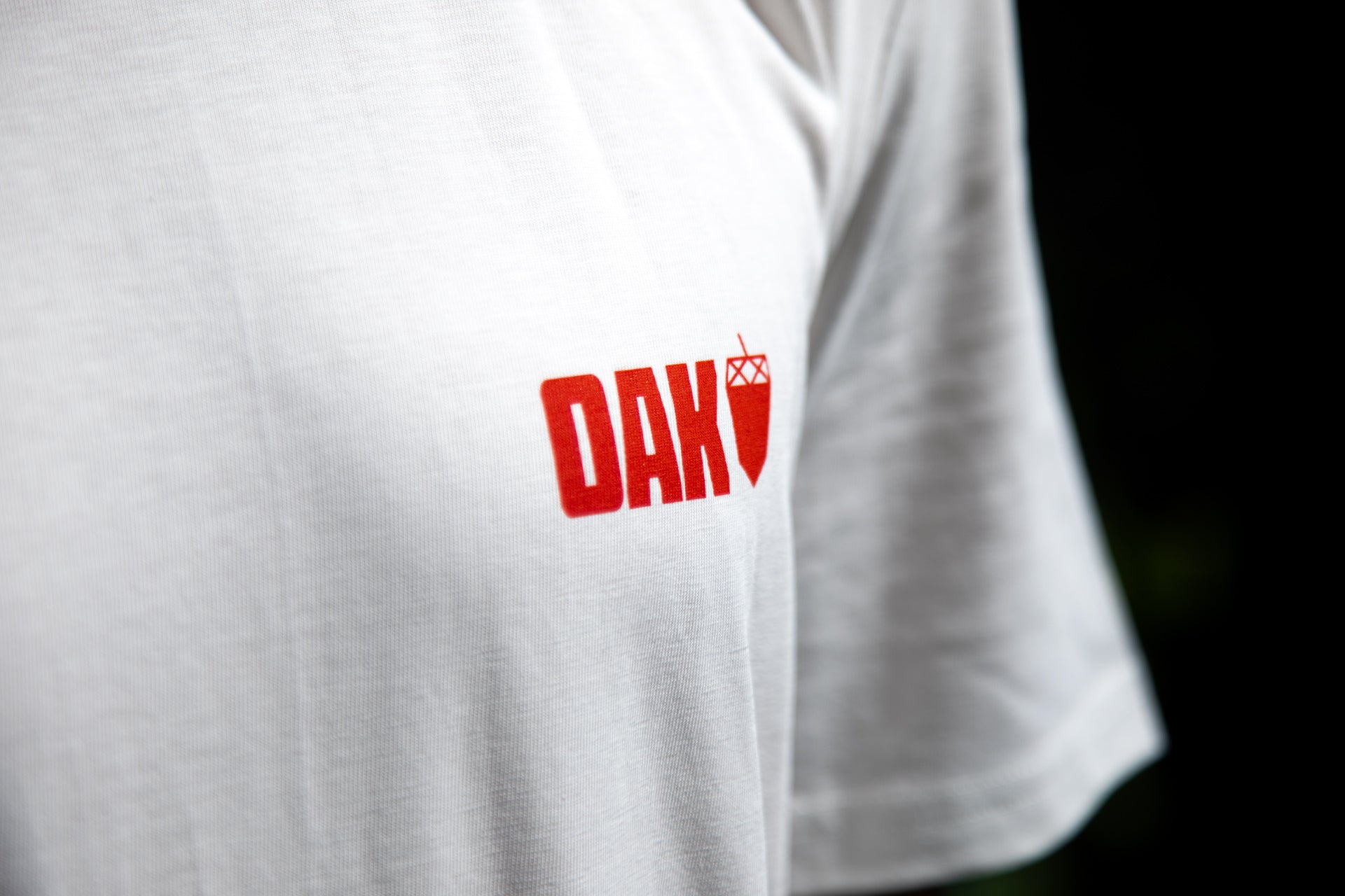 Close-up of a white OAK T-shirt with red logo and graphic detail on chest, showing fabric texture.
