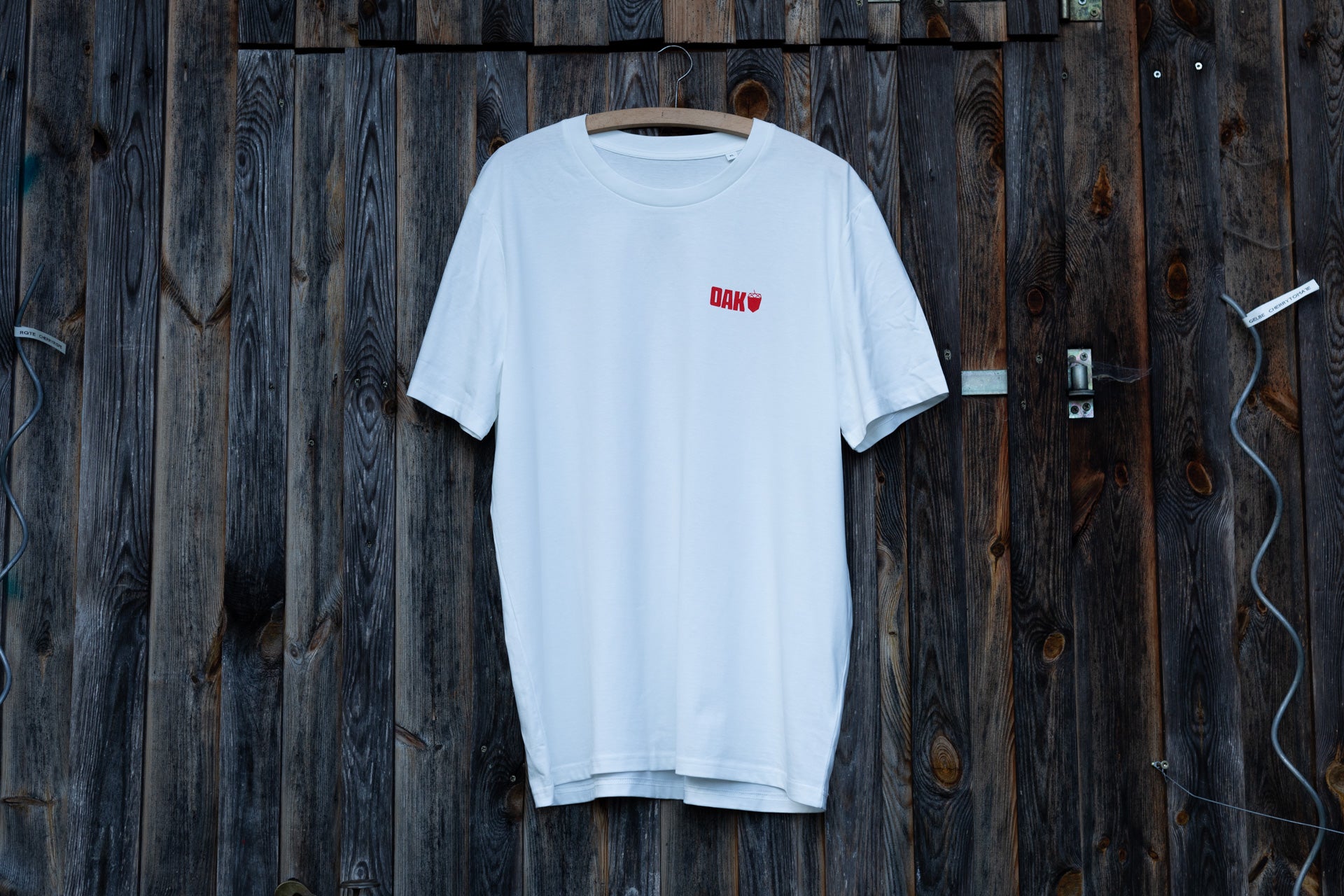 White short-sleeve T-shirt on wooden hanger against dark wood wall with small red OAK logo on chest.