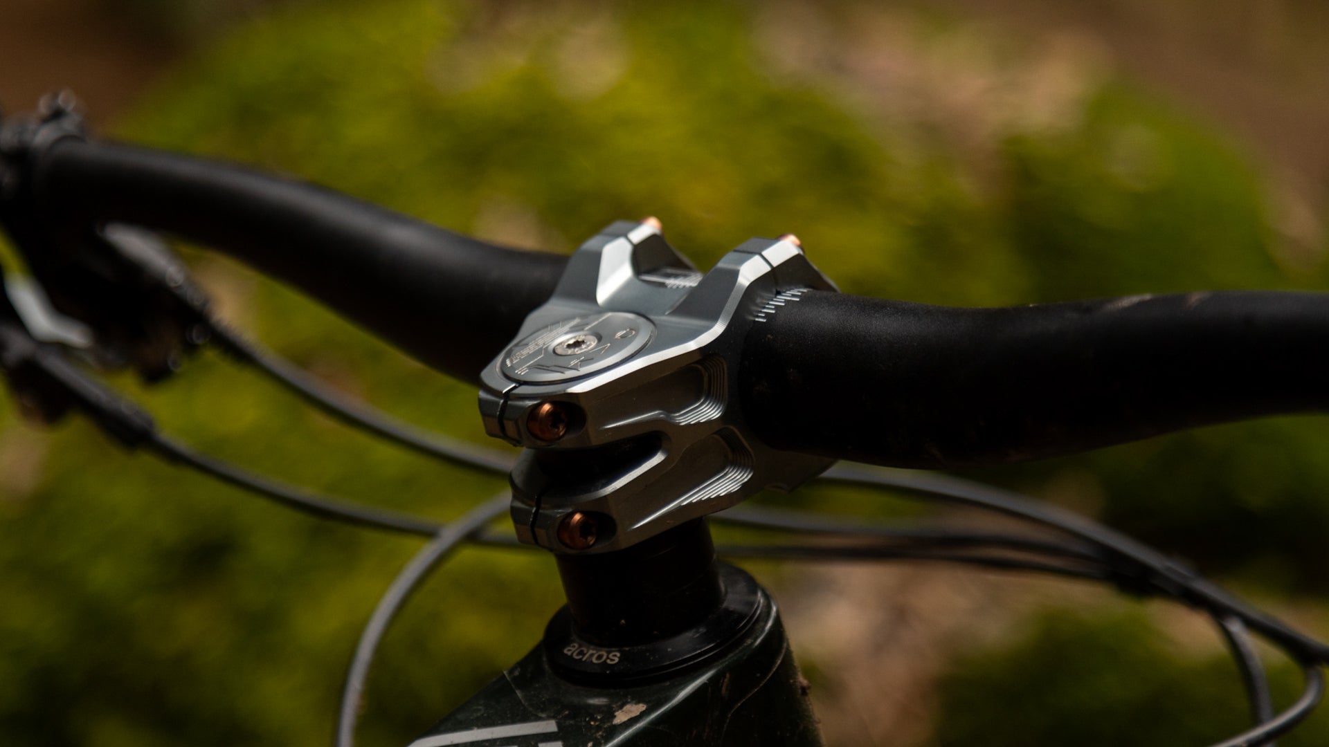 Close-up of a mountain bike handlebar with silver stem and black grips, cables visible, outdoor setting.