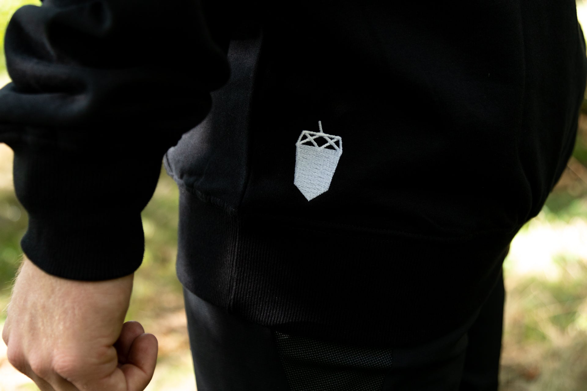 Black sweater with white acorn logo in natural light with green foliage backdrop.
