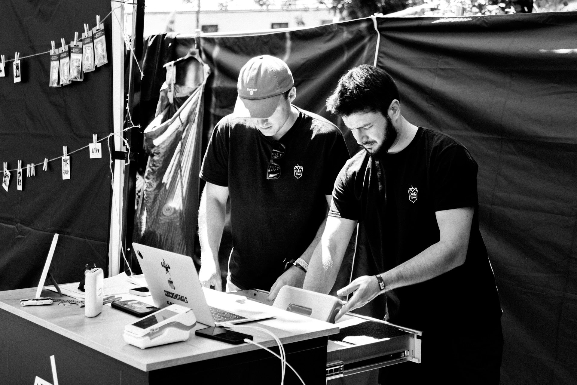 Two men in black OAK TEAM t-shirts at a market table with a laptop, payment terminal, and CHICKENTRAILS stickers.