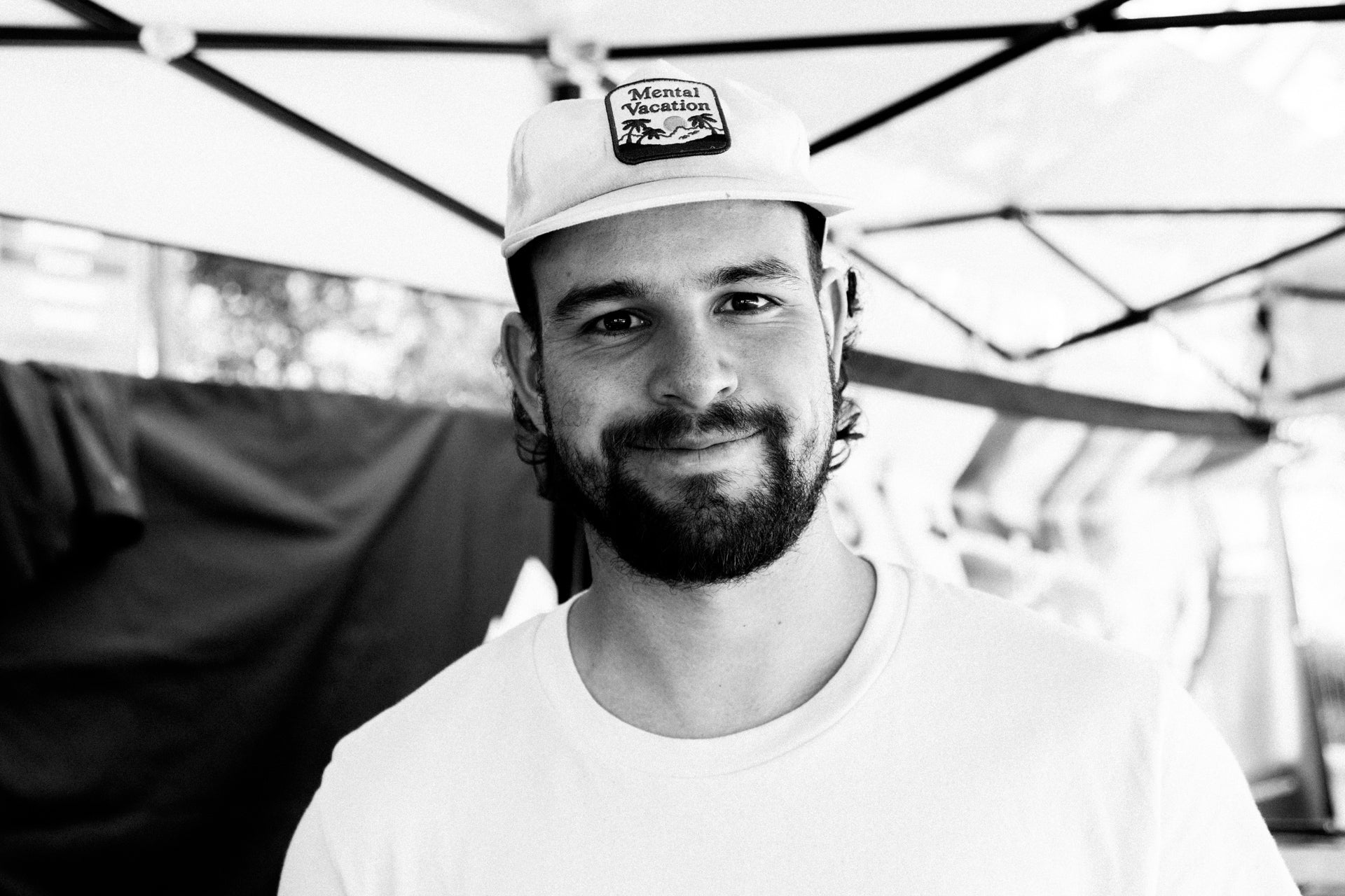 Bearded man in white t-shirt and 'Mental Vacation' cap smiling under a fabric canopy.